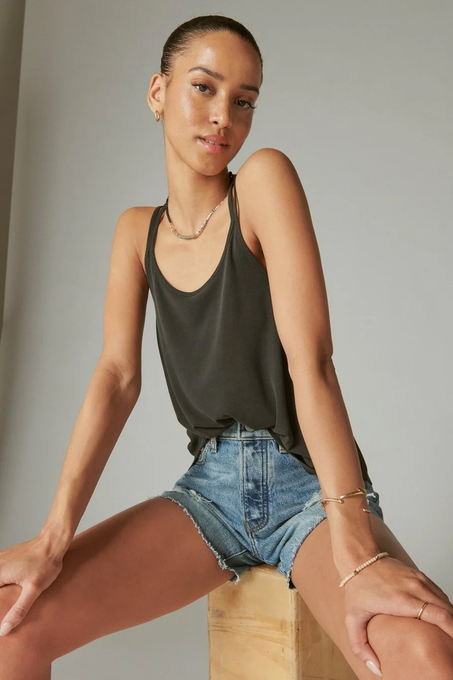 A young woman with fair skin and dark hair tied back, sitting on a wooden block against a plain light-colored wall. She is wearing a black tank top, denim shorts, and various jewelry including necklaces, bracelets, and rings. She is staring at the camera with a relaxed expression.
