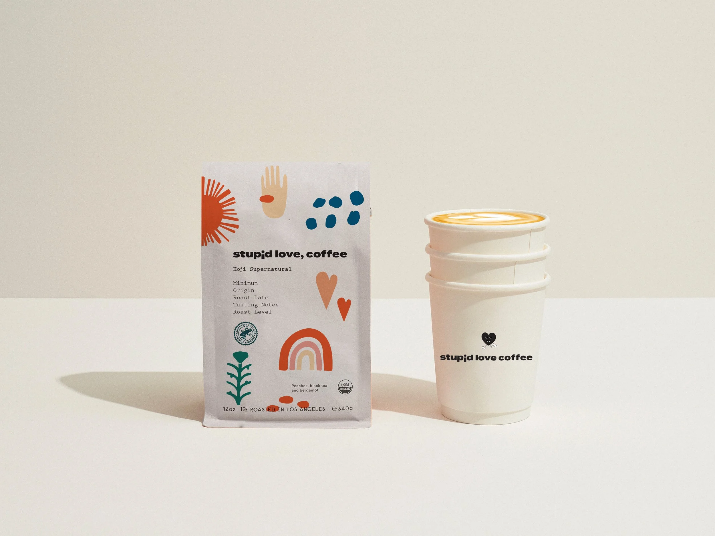 Three stacked white coffee cups with the 'stupid love coffee' logo, next to a bag of coffee beans with colorful abstract designs and text, on a white surface with a shadow.