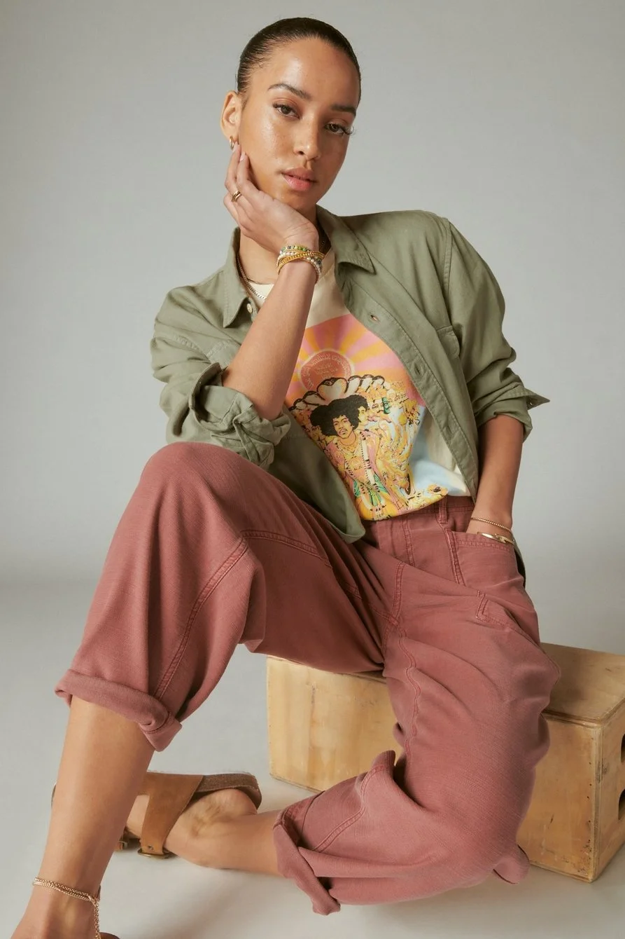 A woman wearing a graphic t-shirt, olive green jacket, and rust-colored pants, sitting on a wooden box against a plain background.