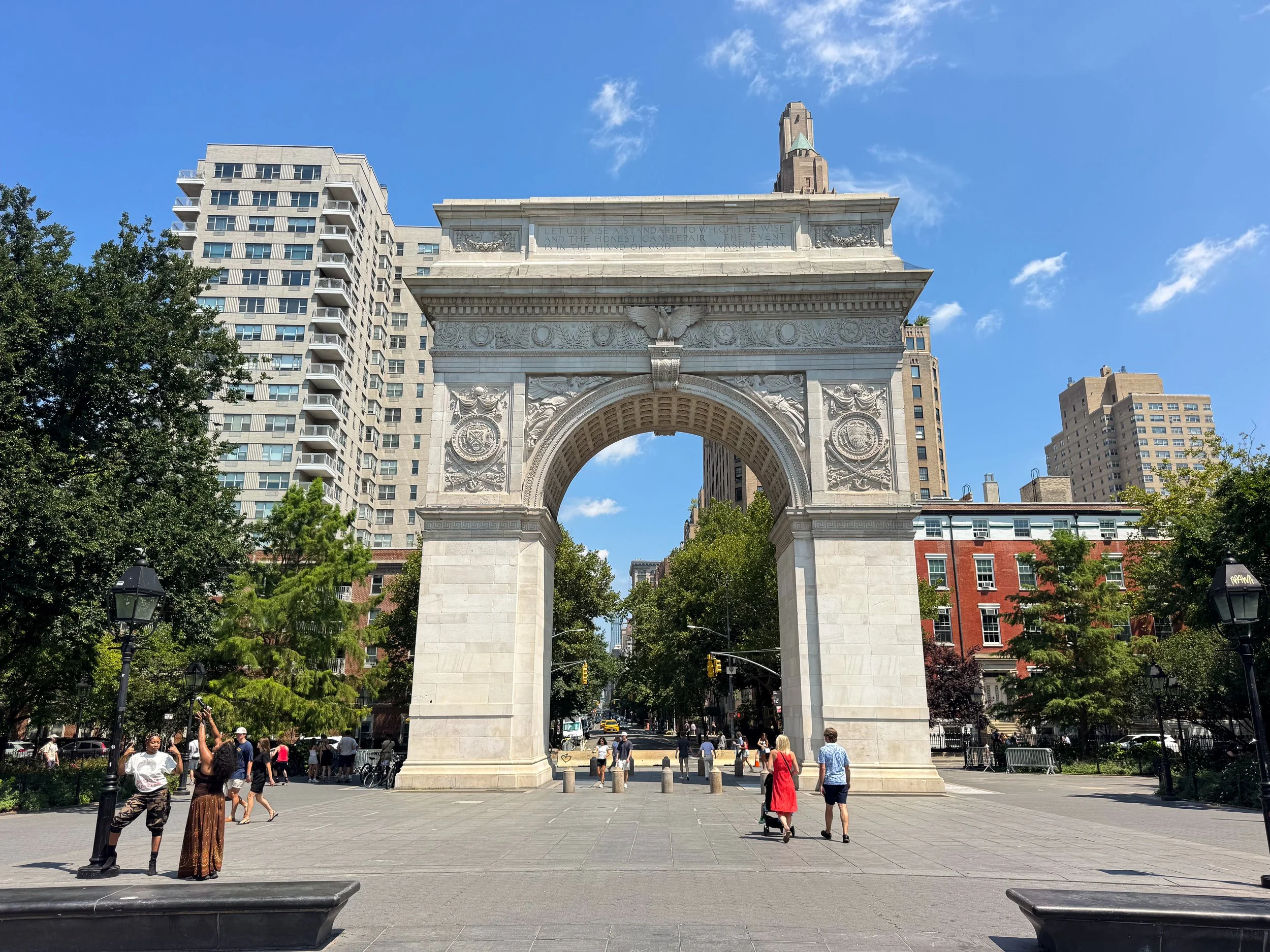 Washington Square Arch rising in white marble above a wide plaza with visitors strolling past shade trees and apartment buildings