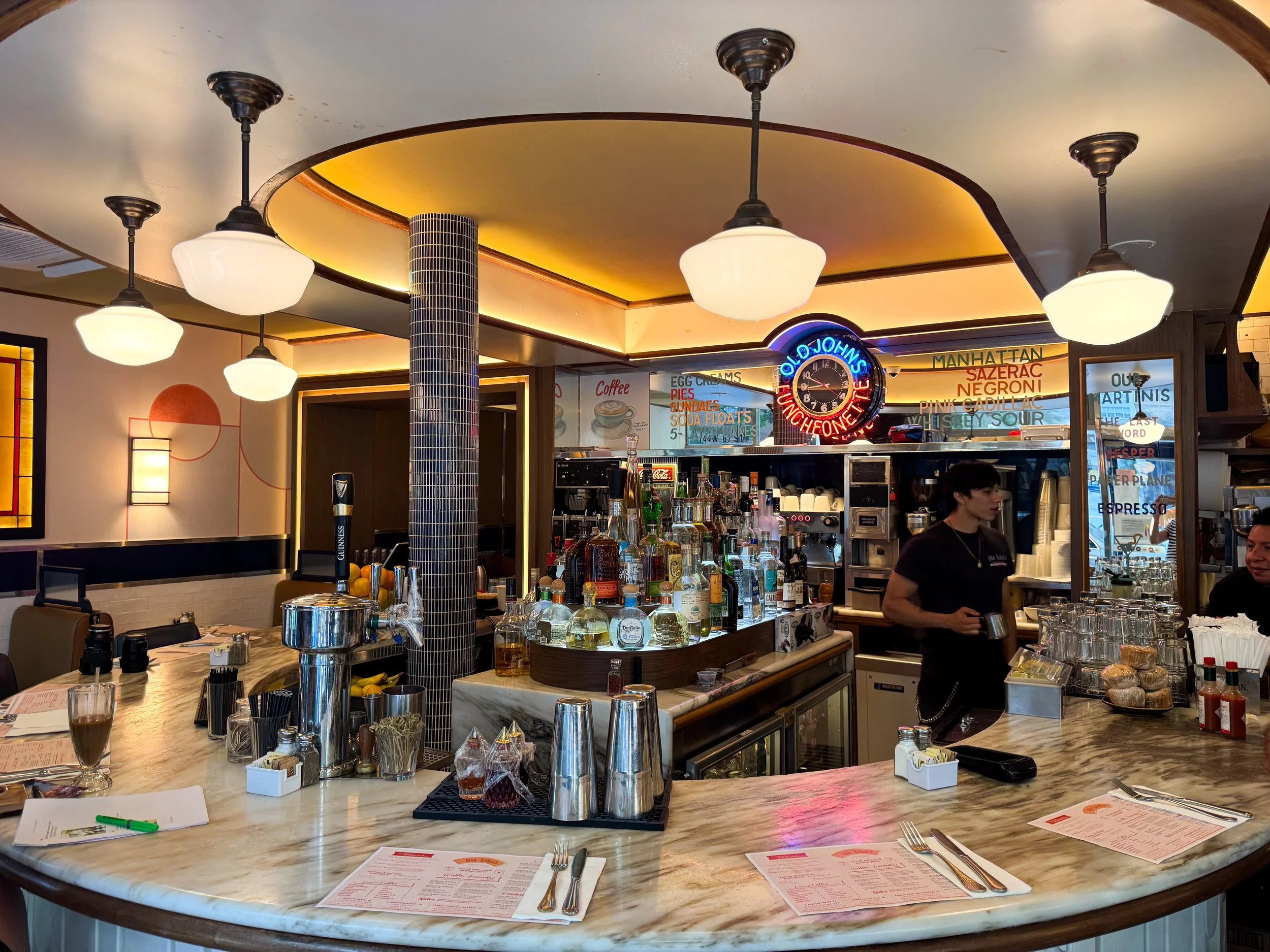 Curved marble counter beneath glowing pendant lights inside an all night diner with a neon clock and rows of liquor bottles