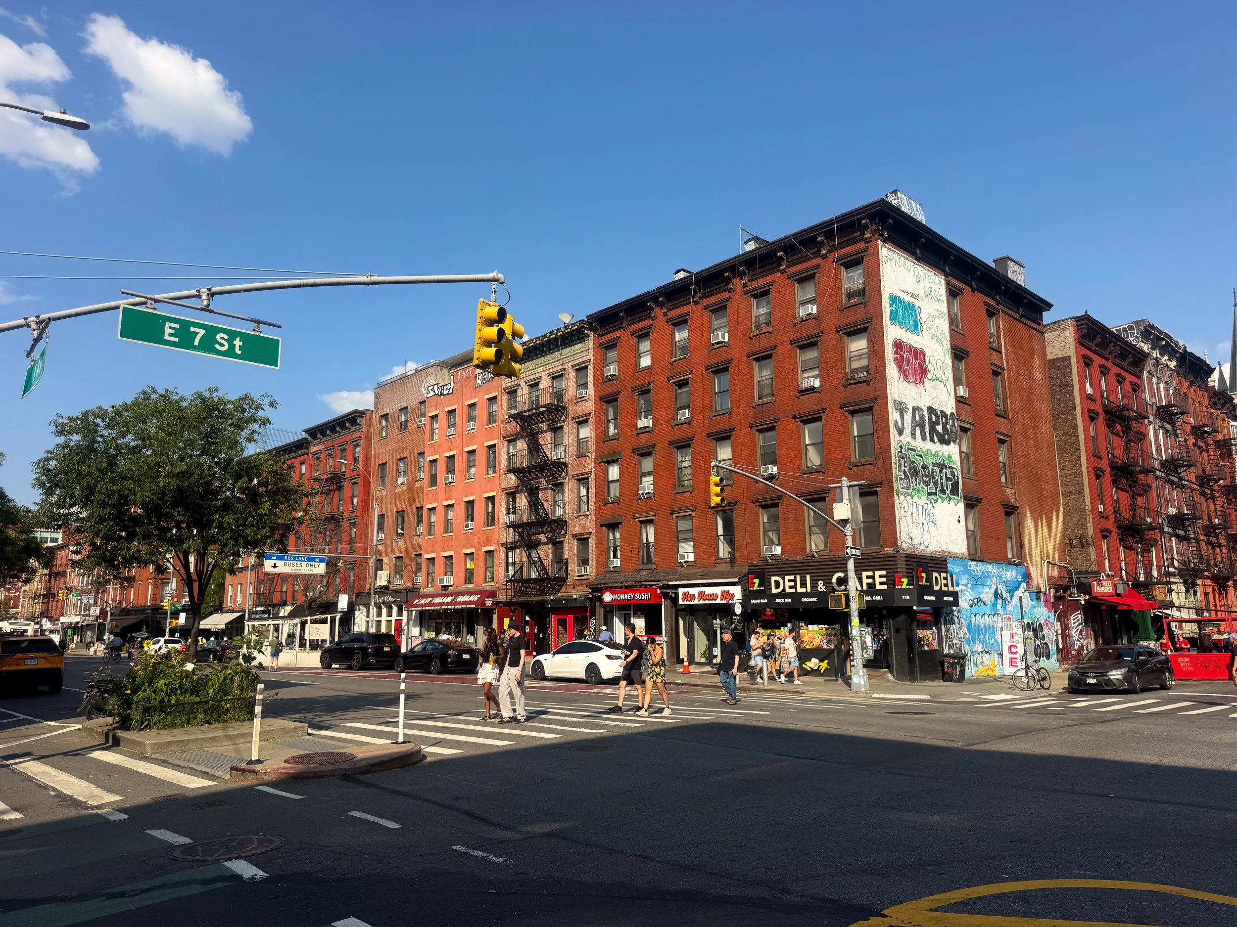 Red brick tenements and a graffiti covered wall above a corner deli in the East Village as pedestrians cross the intersection