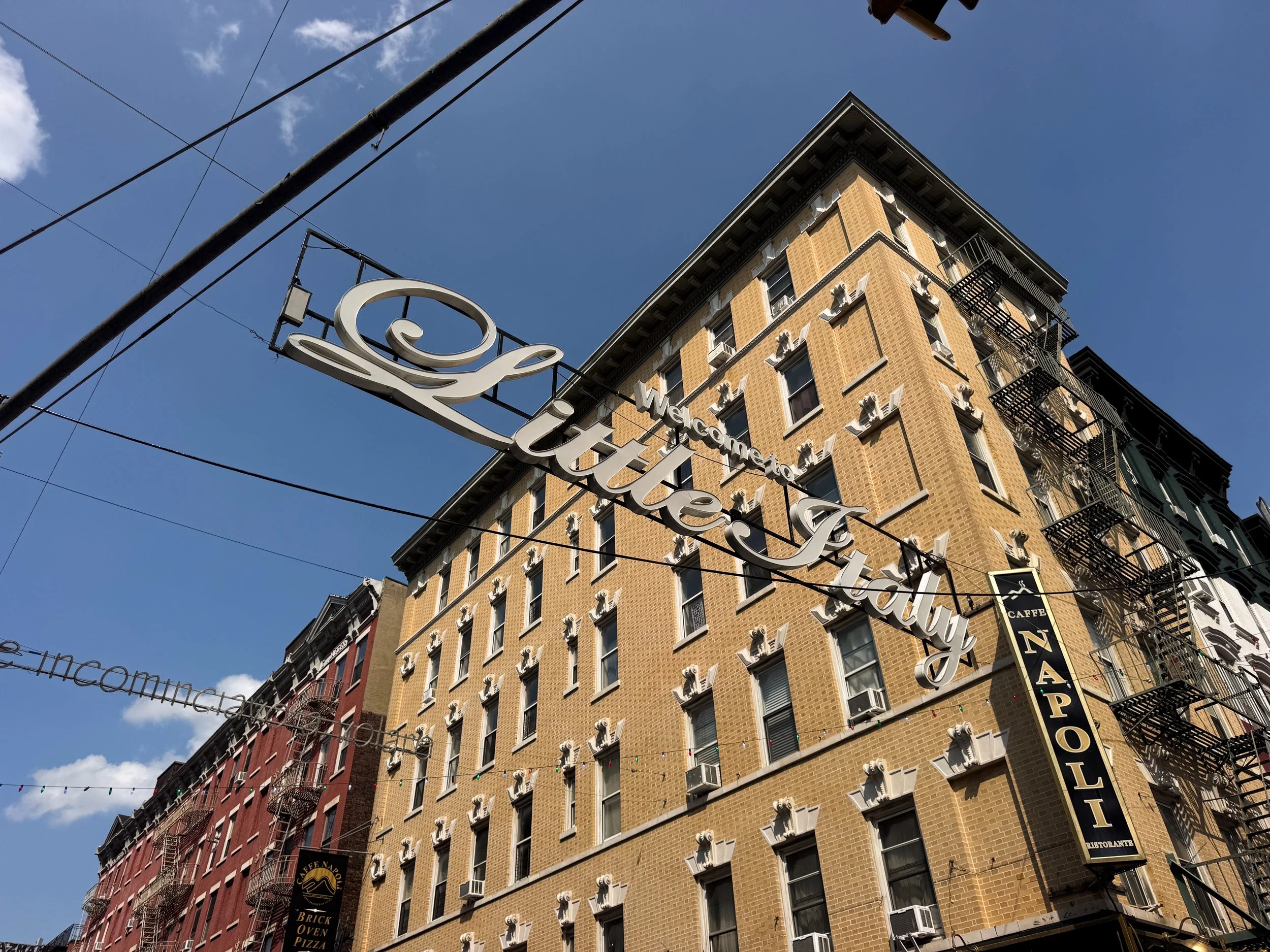 Script sign welcoming visitors to Little Italy strung above the street beside a yellow brick tenement and a Caffe Napoli sign