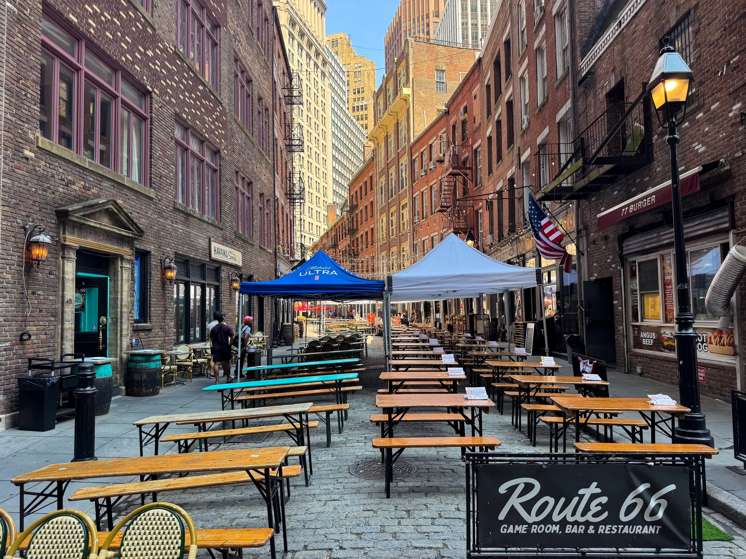 Cobblestone pedestrian street lined with wooden picnic benches, canopies and historic brick buildings set for outdoor dining beneath a street lamp