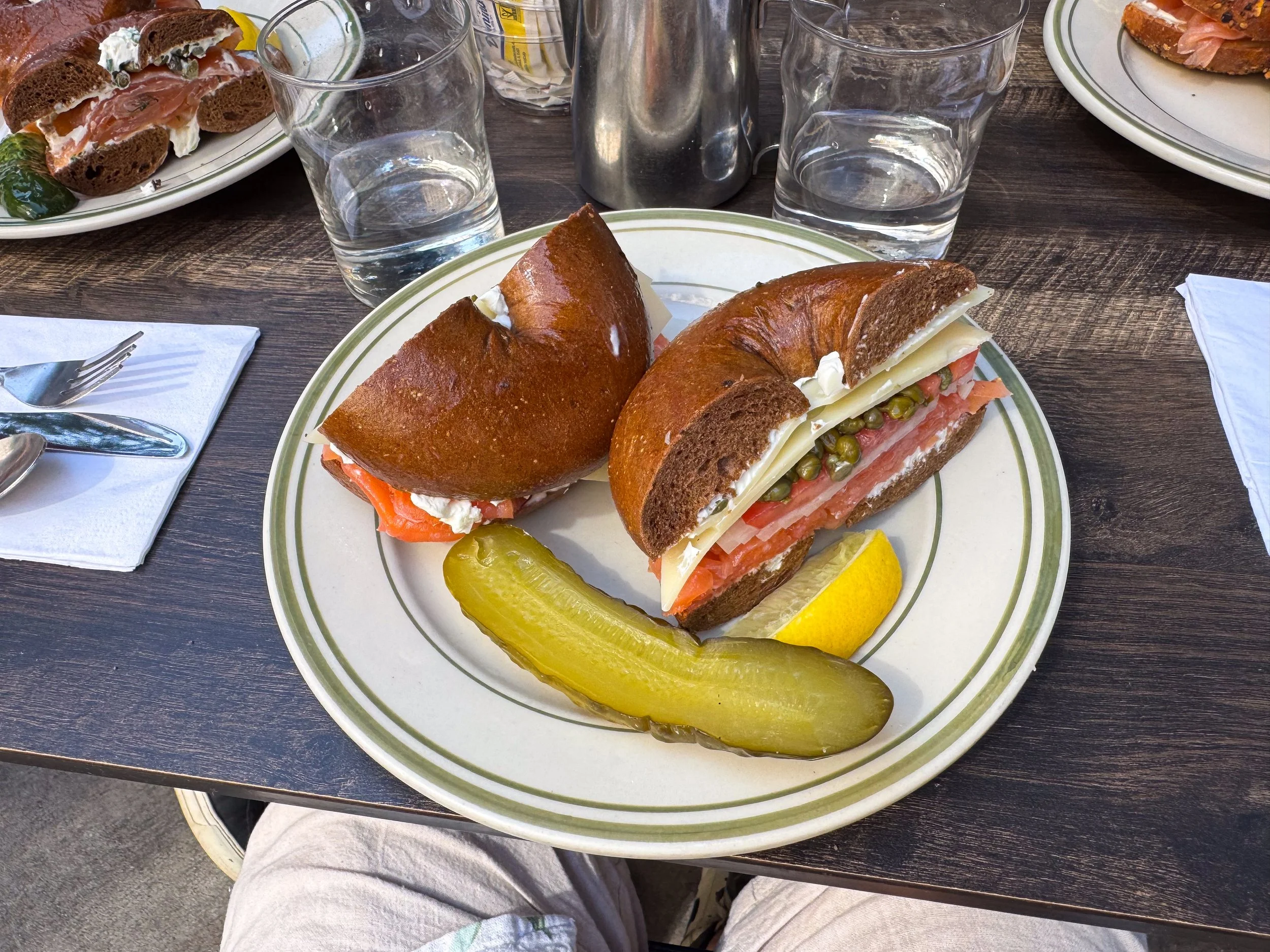 Pumpernickel bagel with lox, cream cheese, capers and a pickle spear plated at a diner table with more bagels nearby