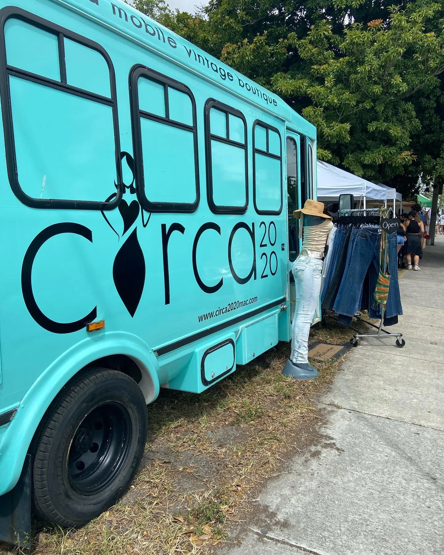 A bright turquoise vintage food truck with the words "mobile vintage boutique" and "circa 2020" on the side, parked at an outdoor market. A mannequin wearing a sunhat and light-colored clothing stands outside the truck, next to a rack of jeans and cl