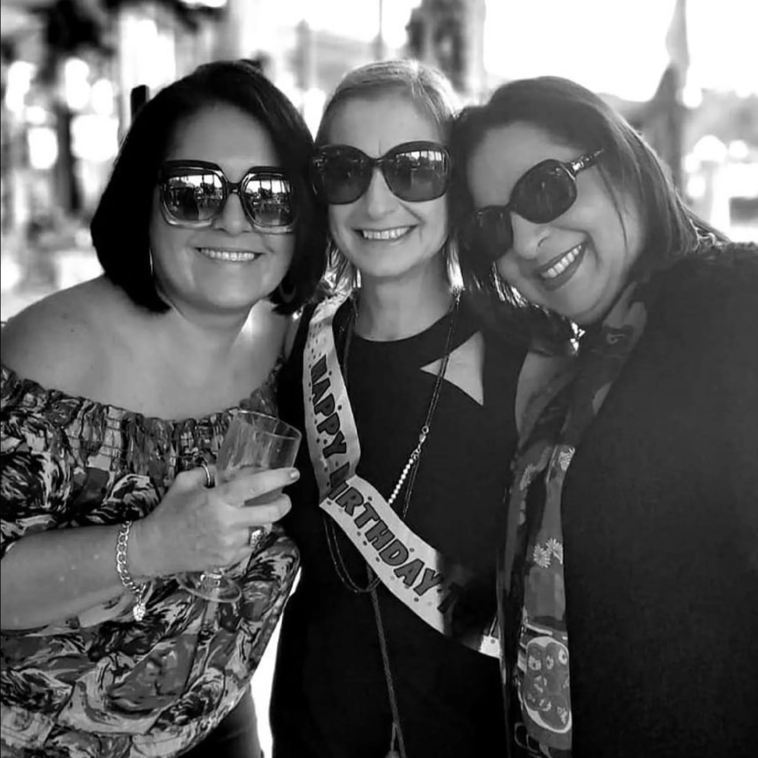 Three women smiling, wearing sunglasses, at a birthday celebration, one woman has a 'Happy Birthday' sash.
