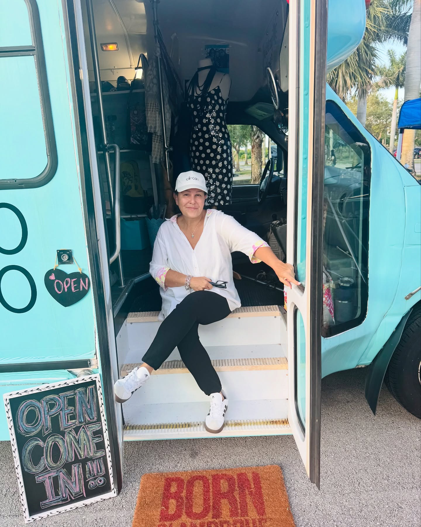 A woman sitting on the steps of a turquoise food truck, smiling at the camera, with a black and white Dressed mannequin inside the truck and signs that say 'Open' and 'Come In' outside.