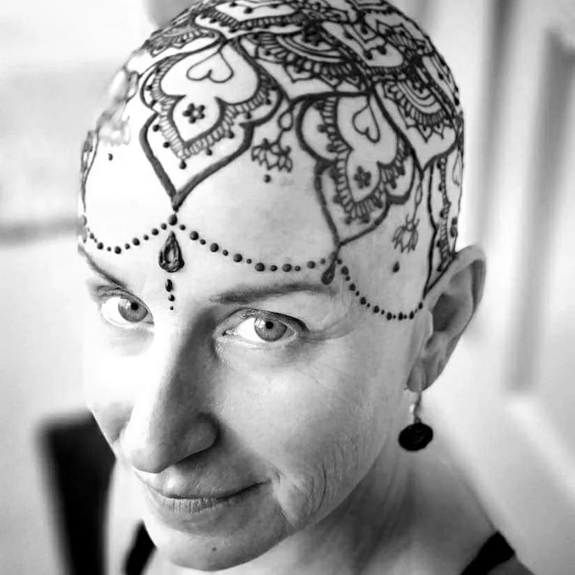 Black and white photo of a woman with a detailed henna or tattoo design on her head. She has earrings and is looking at the camera with a slight smile.