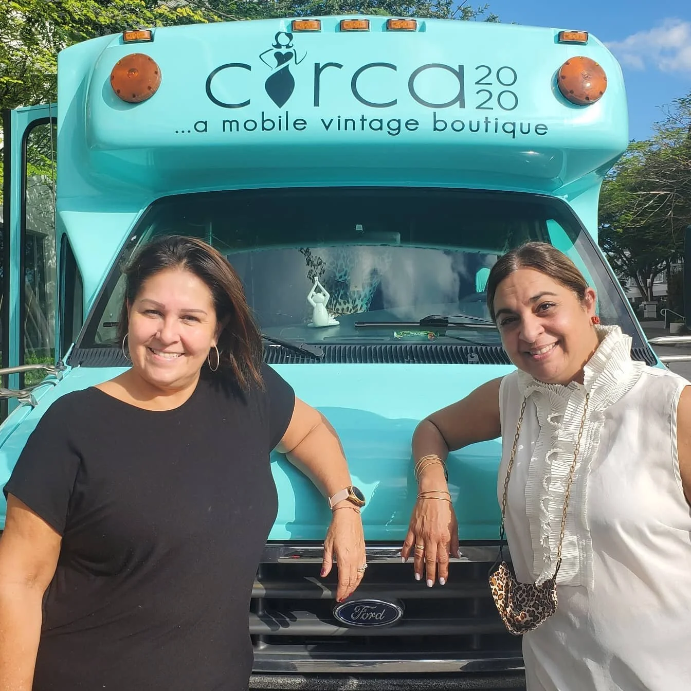 Two women standing in front of a turquoise Ford food truck with a sign 'Cora 2020, a mobile vintage boutique.' They are smiling and posing for the photo.