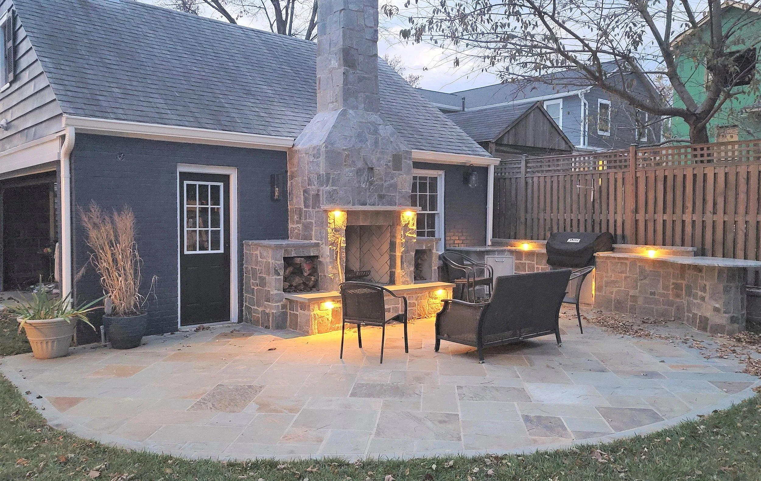 Backyard patio area with an outdoor fireplace, patio furniture, potted plants, and lighting, enclosed by a wooden fence.