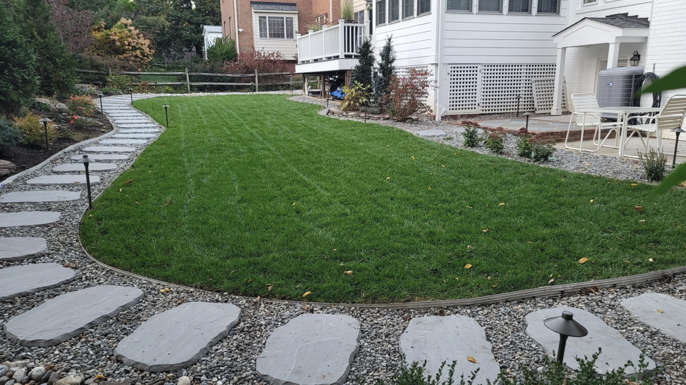 A backyard with a green grassy lawn surrounded by a curved stepping stone pathway which hides stormwater drainage, small garden lights, and plants along the edges.