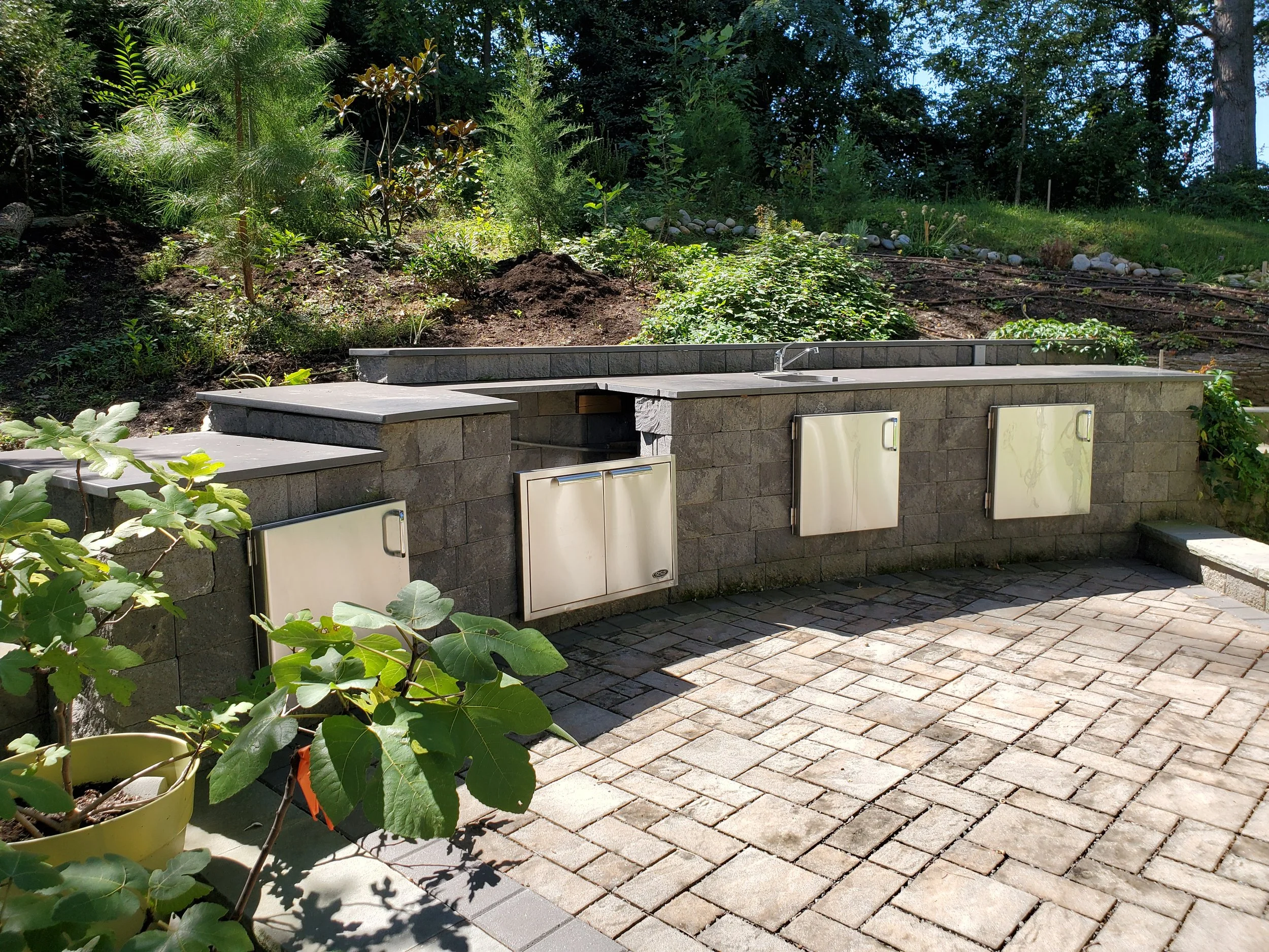 Outdoor kitchen with a curved stone counter, stainless steel cabinets, a small sink, surrounded by plants and brick pavers.