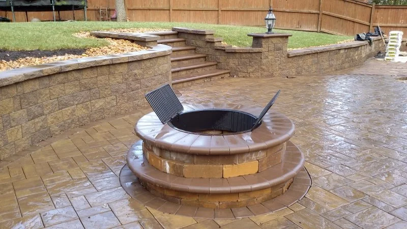 A circular brick fire pit with a black grill and a metal fire poker, surrounded by wet stone patio, with stairs and a grassy backyard in the background.