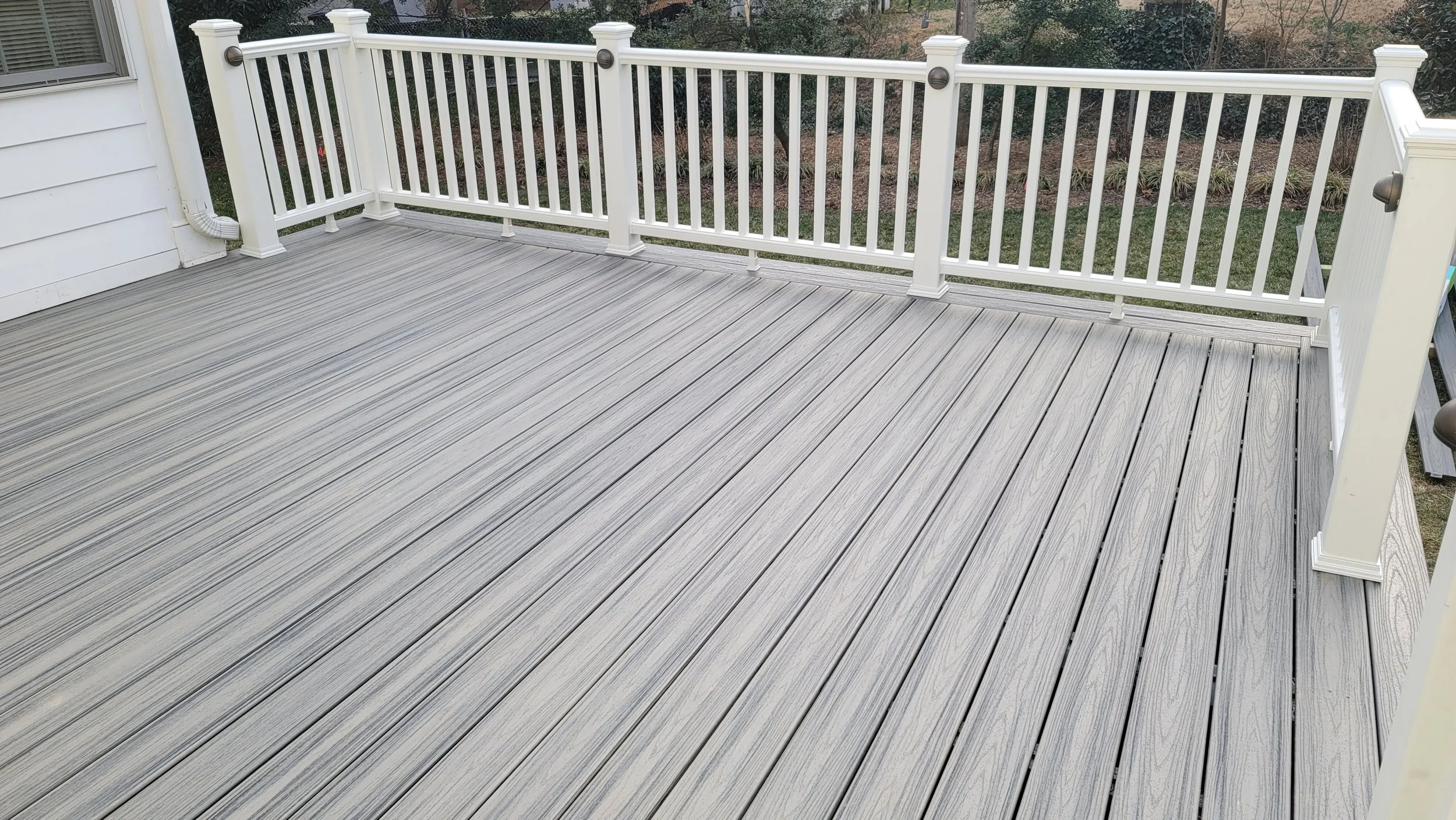 A spacious outdoor deck with light gray wooden planks, enclosed by white railing with cylindrical posts, attached to a white house with siding, and surrounded by trees and bushes in the background.
