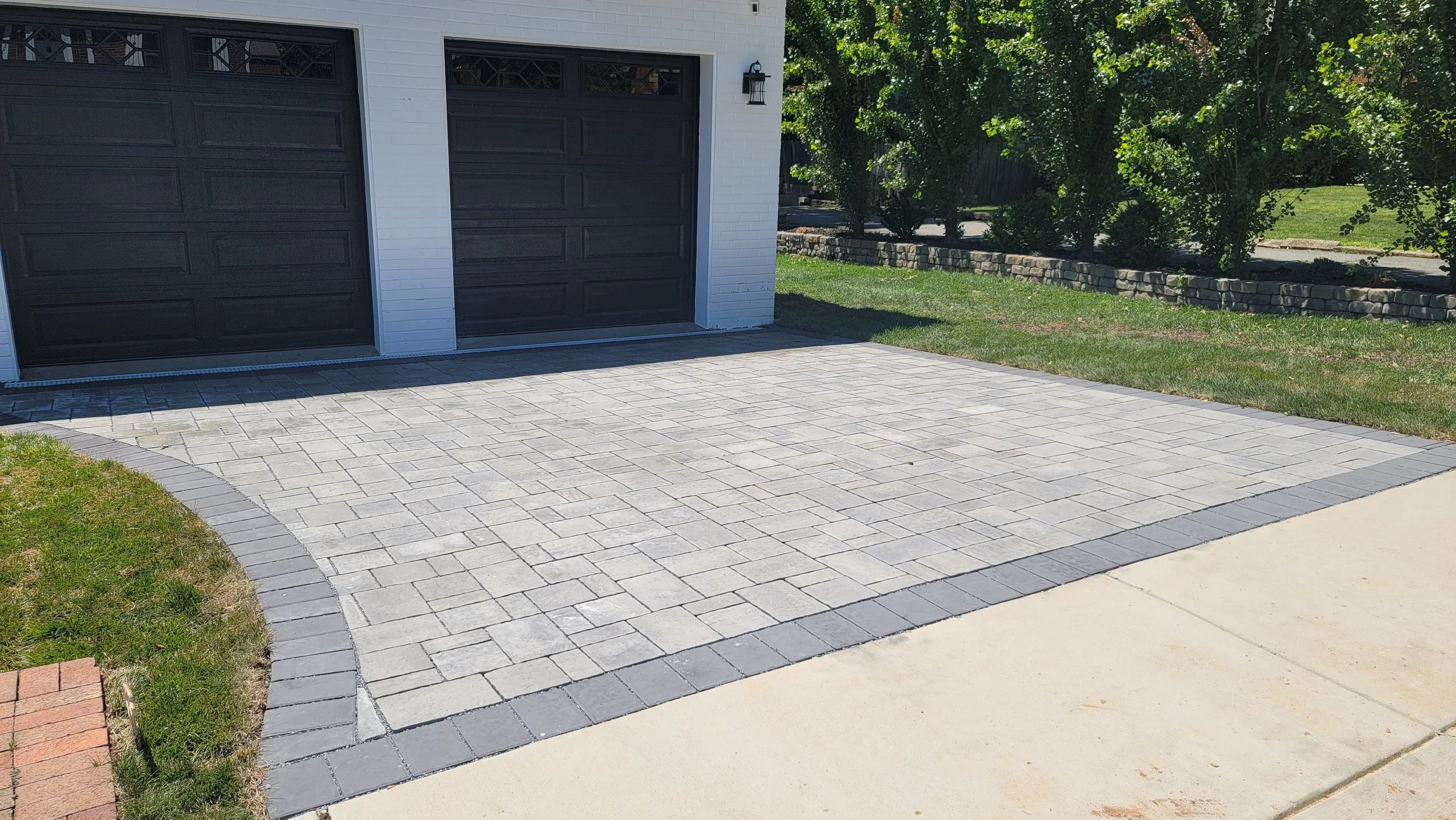 A paved driveway with light-colored bricks bordered by darker bricks, adjacent to a concrete sidewalk, next to a white brick garage with black doors and a small outdoor light, with a grassy yard and bushes in the background.