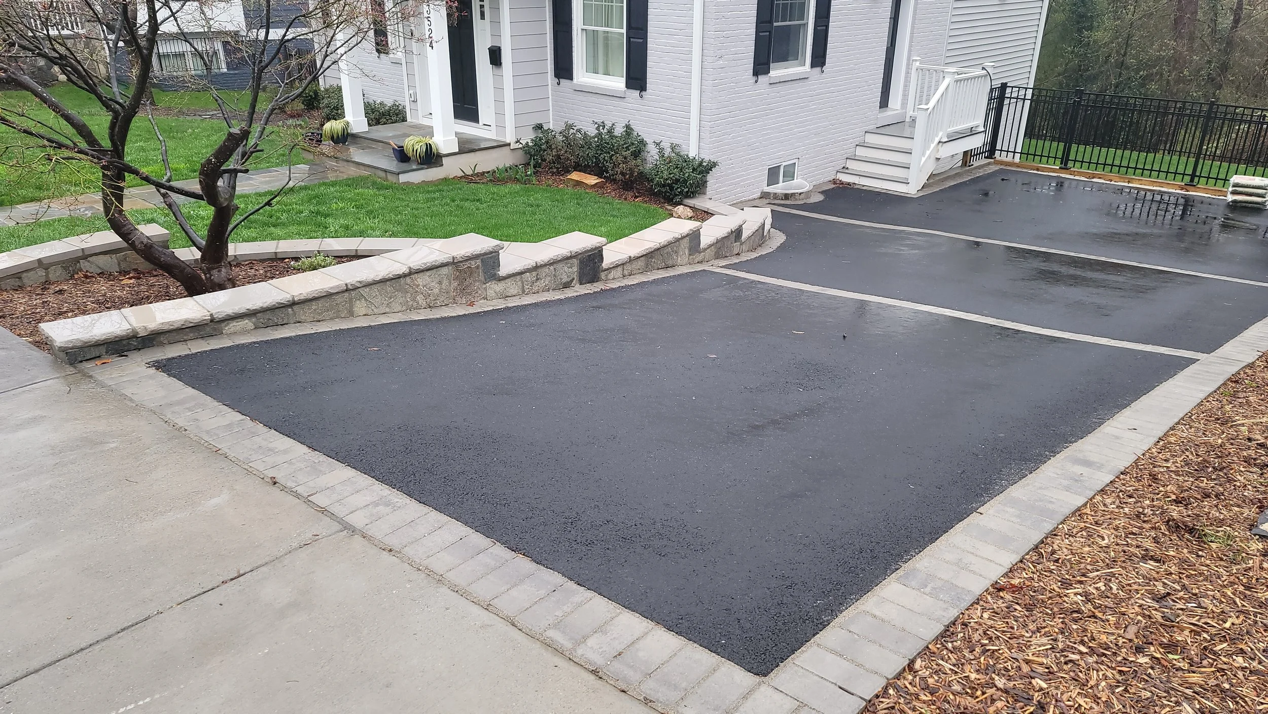 Empty newly paved parking spaces in front of a residential house with a garden, tree, and sidewalk.