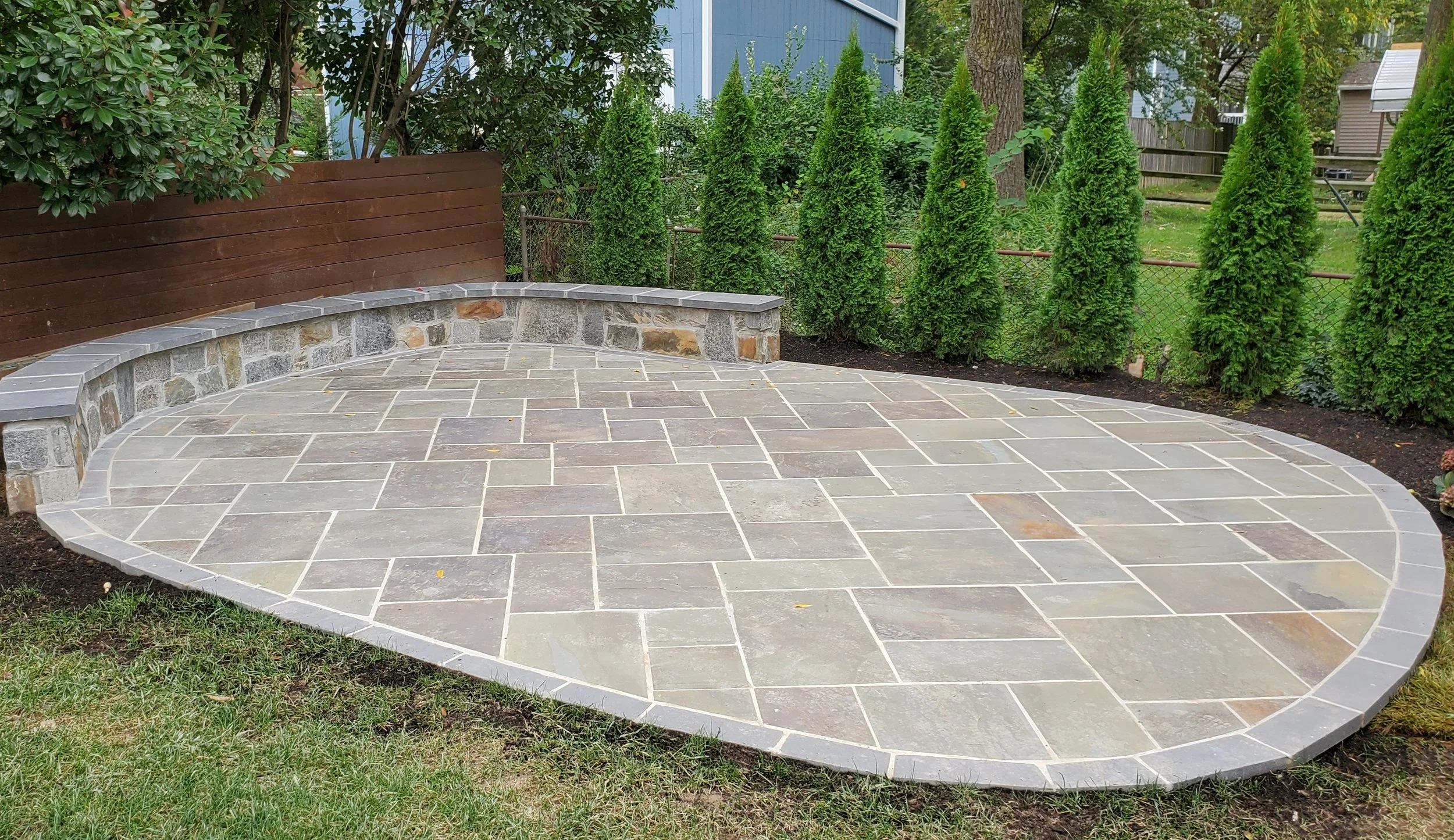 Curved backyard patio with stone tiles, bordered by gray bricks, surrounded by green grass, shrubs, and tall evergreen trees, with a blue building in the background.