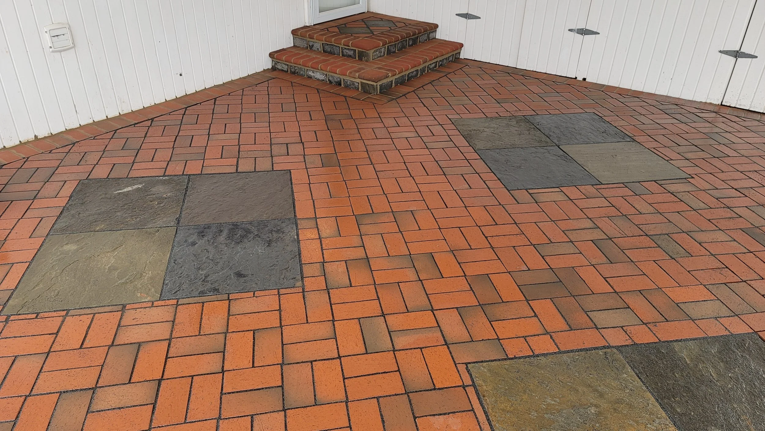 Exterior brick patio with patterned red and gray paving stones and small brick stairs leading up to a door.