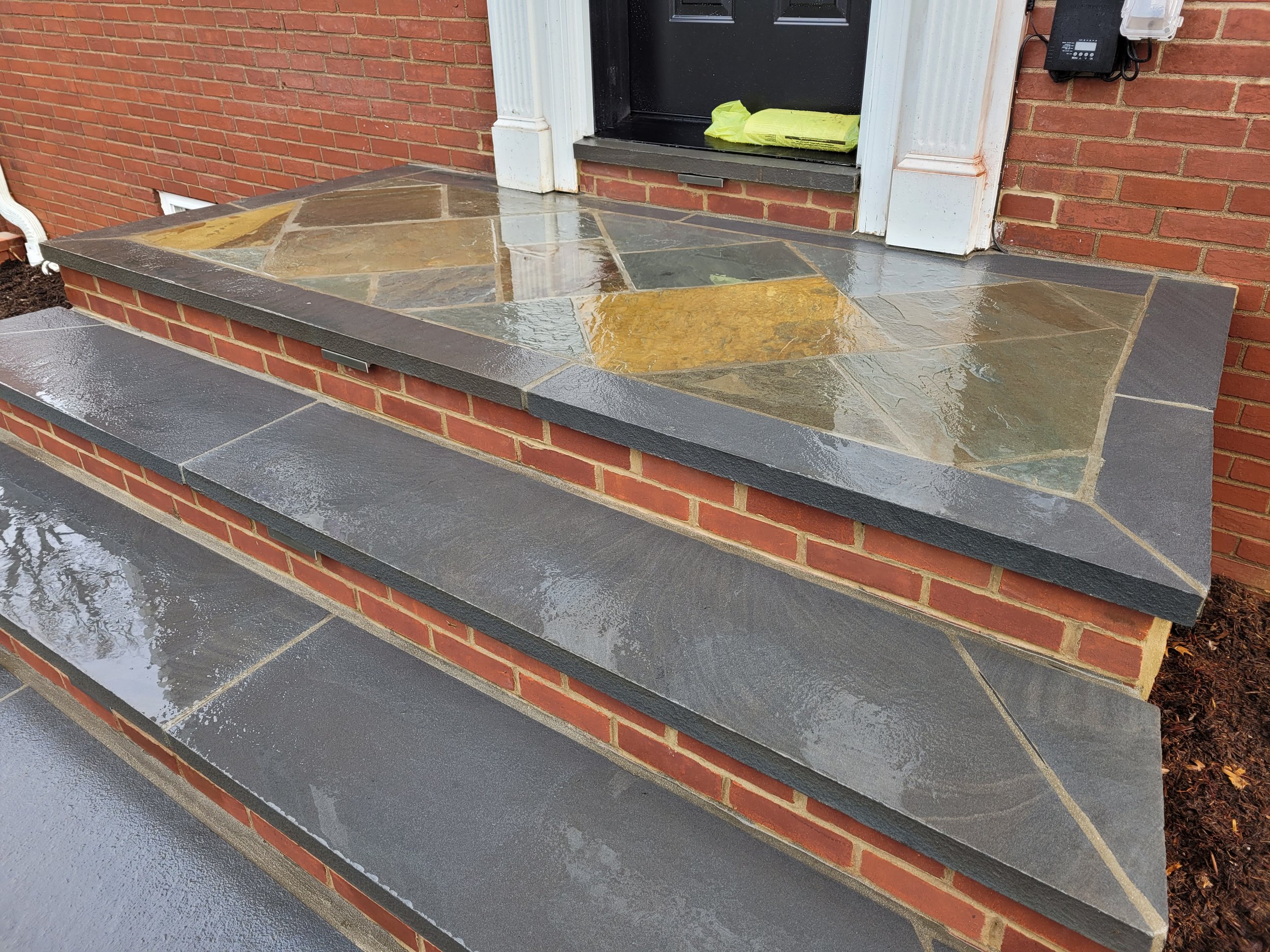 Newly constructed front porch with brick foundation, stone steps, and a slate tile landing in front of a black door with white trim.