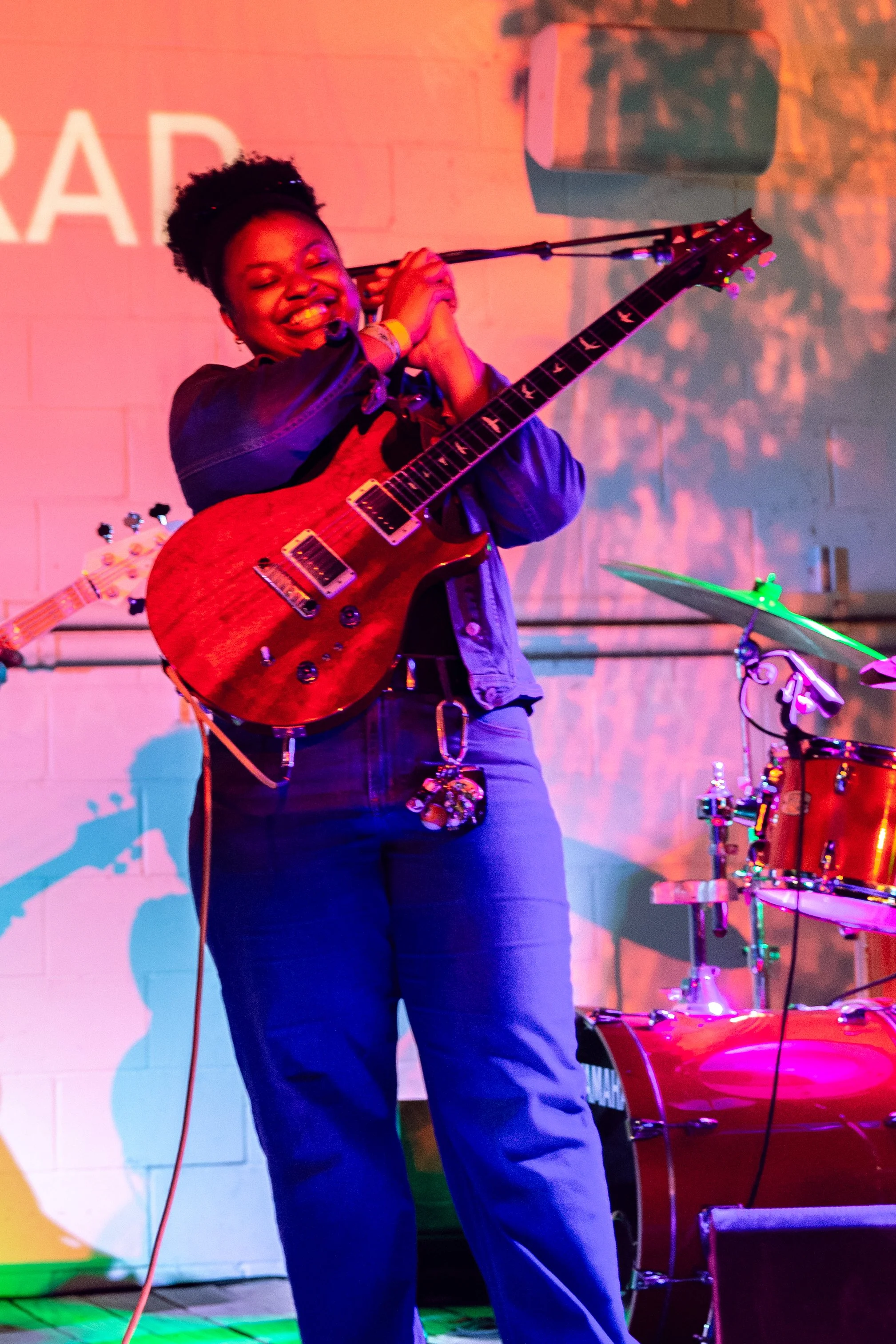 A woman playing an electric guitar in a dimly lit room with purple and pink lighting, shelves with records and artwork on the wall behind her.
