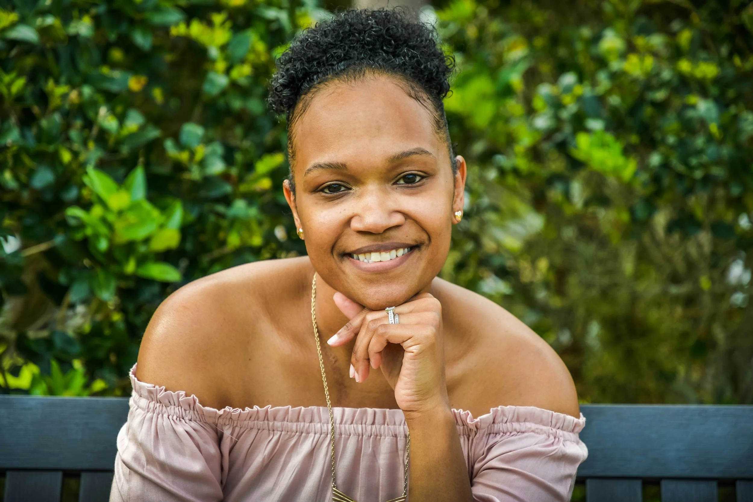 Close-up of a young woman with dark, curly hair styled in a bun, smiling with her hand resting on her chin, wearing an off-shoulder pink top, sitting on a black bench outdoors with green foliage in the background.