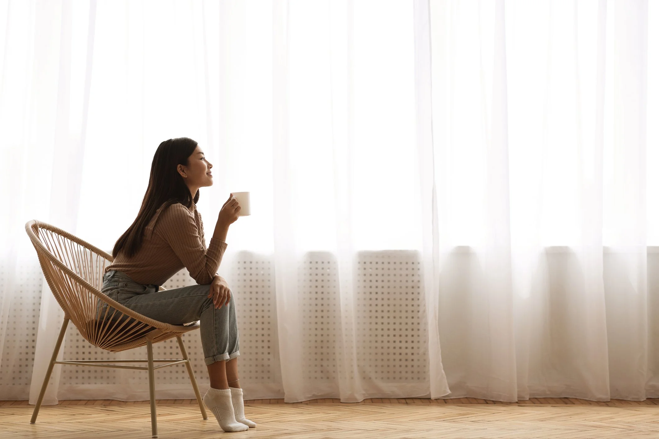 Woman sitting on a wicker chair, holding a mug, by a large window with sheer curtains, wearing a ribbed sweater, jeans, and socks, in a bright, minimalistic room.