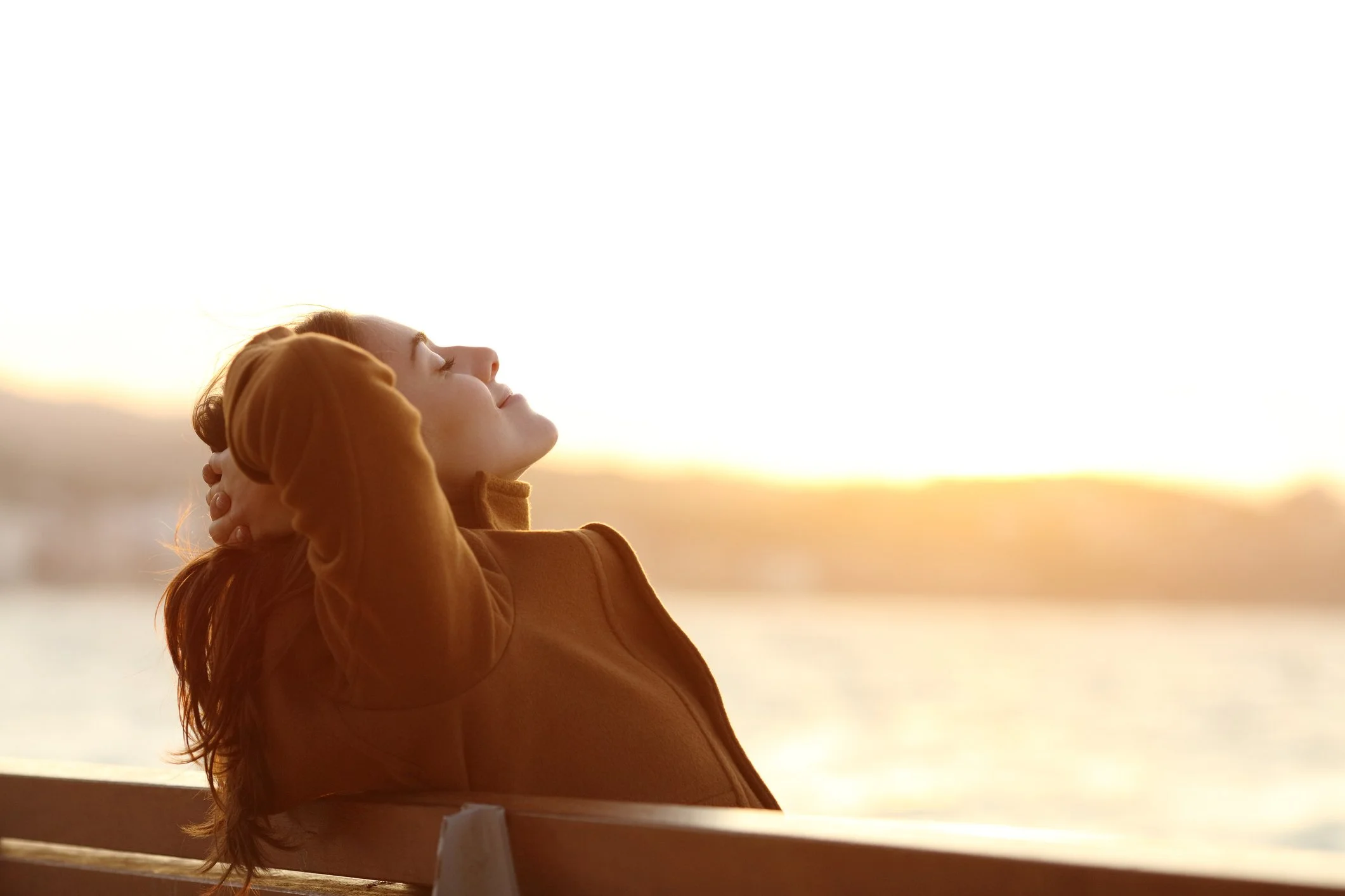 A woman with long hair in a brown jacket leaning back and enjoying the sunset near a body of water.