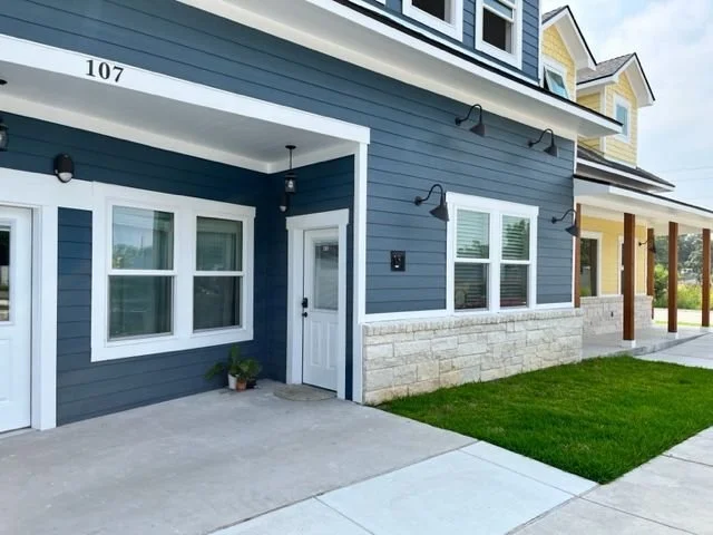 Front porch of a modern blue house with white trim, a white door, three windows, and a small potted plant near the entrance. House number 107 visible above the porch.