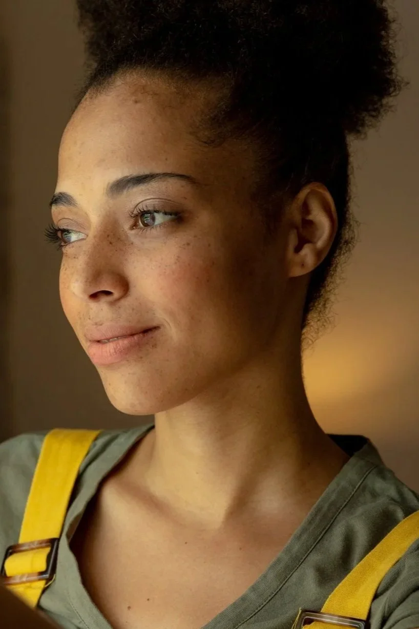 A young woman with dark, curly hair tied back, light brown skin with freckles, and light-colored eyes looking off to the side, wearing a green top with yellow straps.
