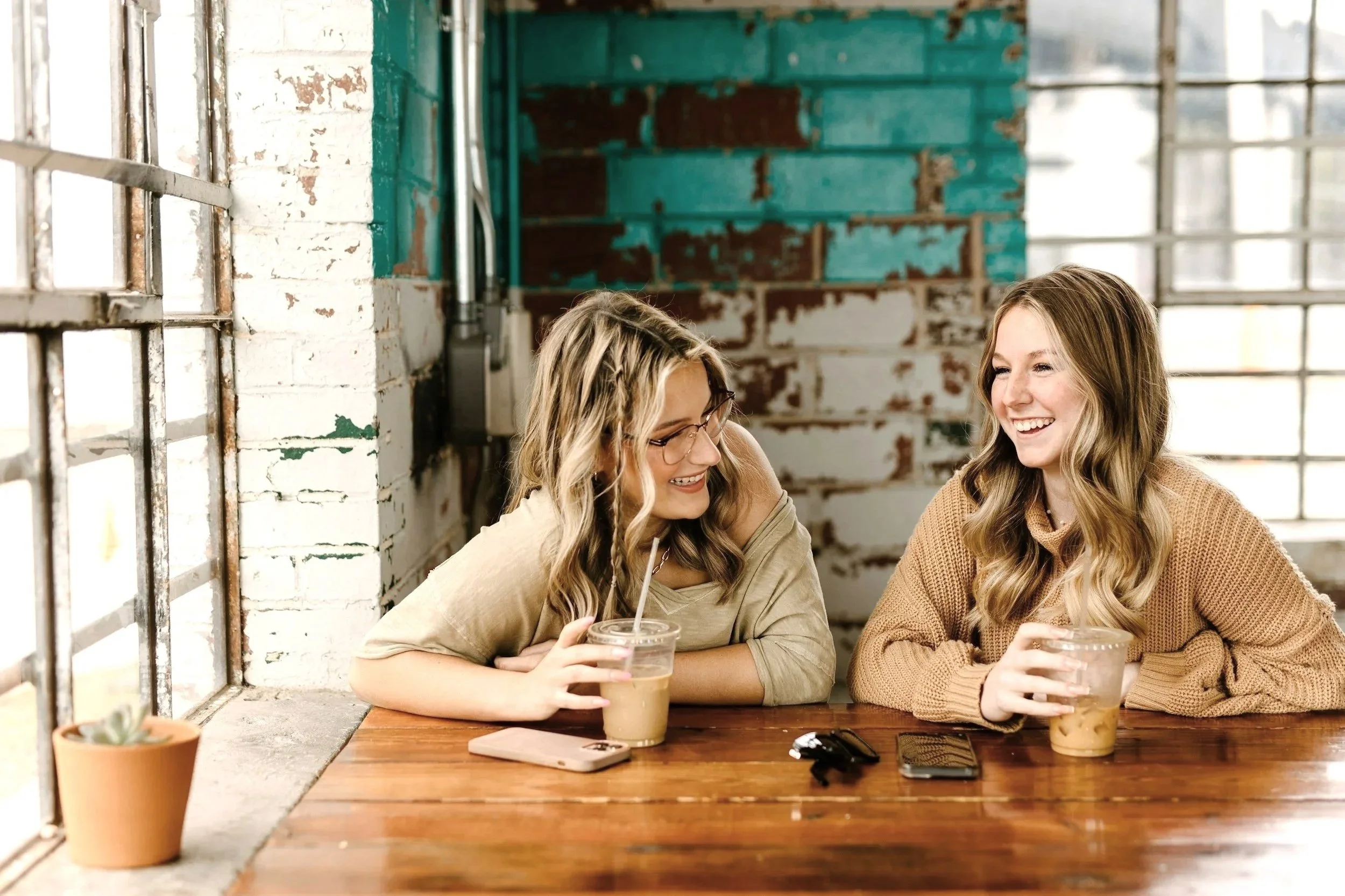 Two women sitting at a wooden table, sharing a laugh, each holding a glass of iced coffee. The woman on the left has long wavy blonde hair and glasses, the woman on the right has long wavy light brown hair. There are two smartphones and a small potted plant on the table, with a rustic, industrial-style background with exposed brick and large windows letting in natural light.