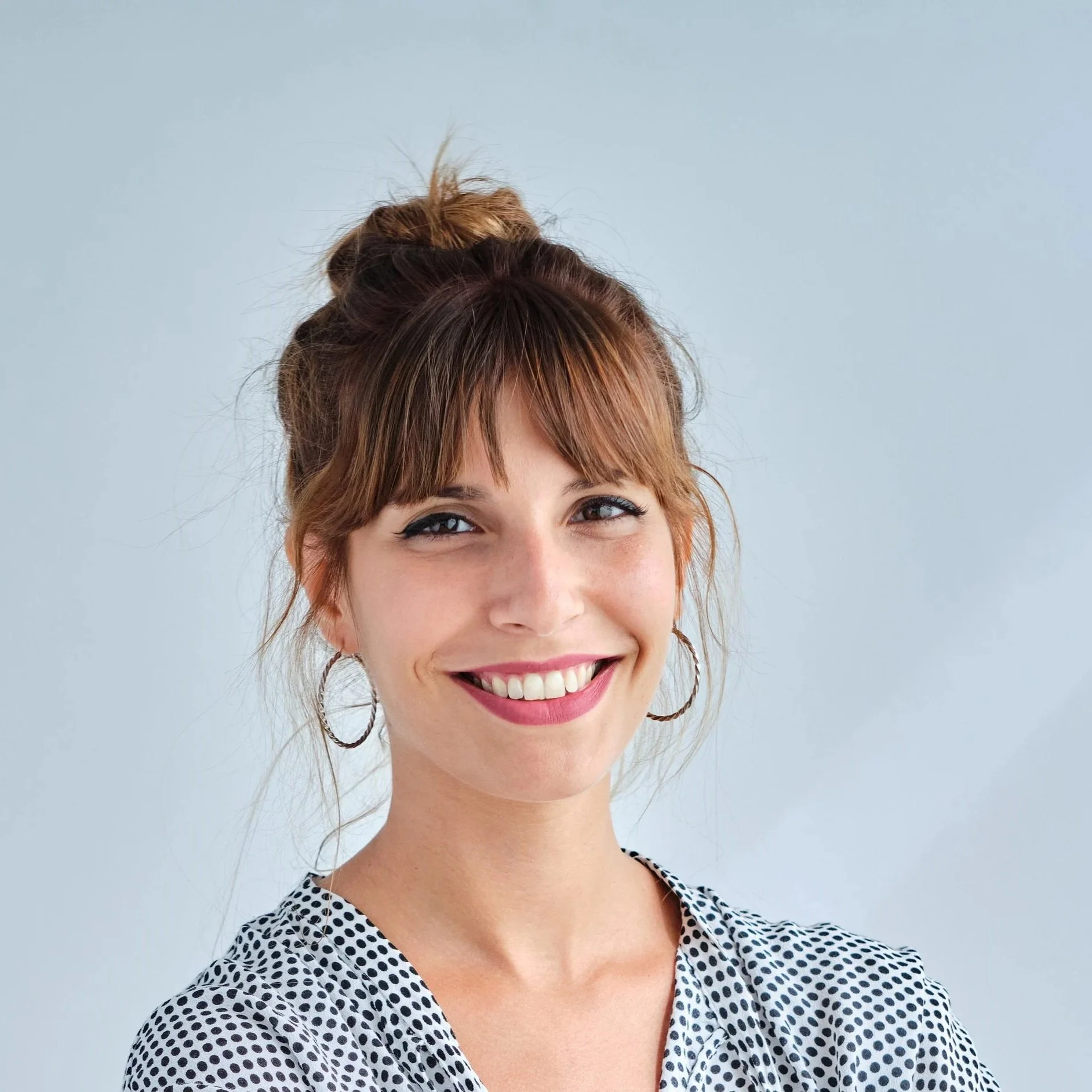 A smiling woman with red hair, wearing hoop earrings and a black and white polka dot blouse, standing against a light gray background.
