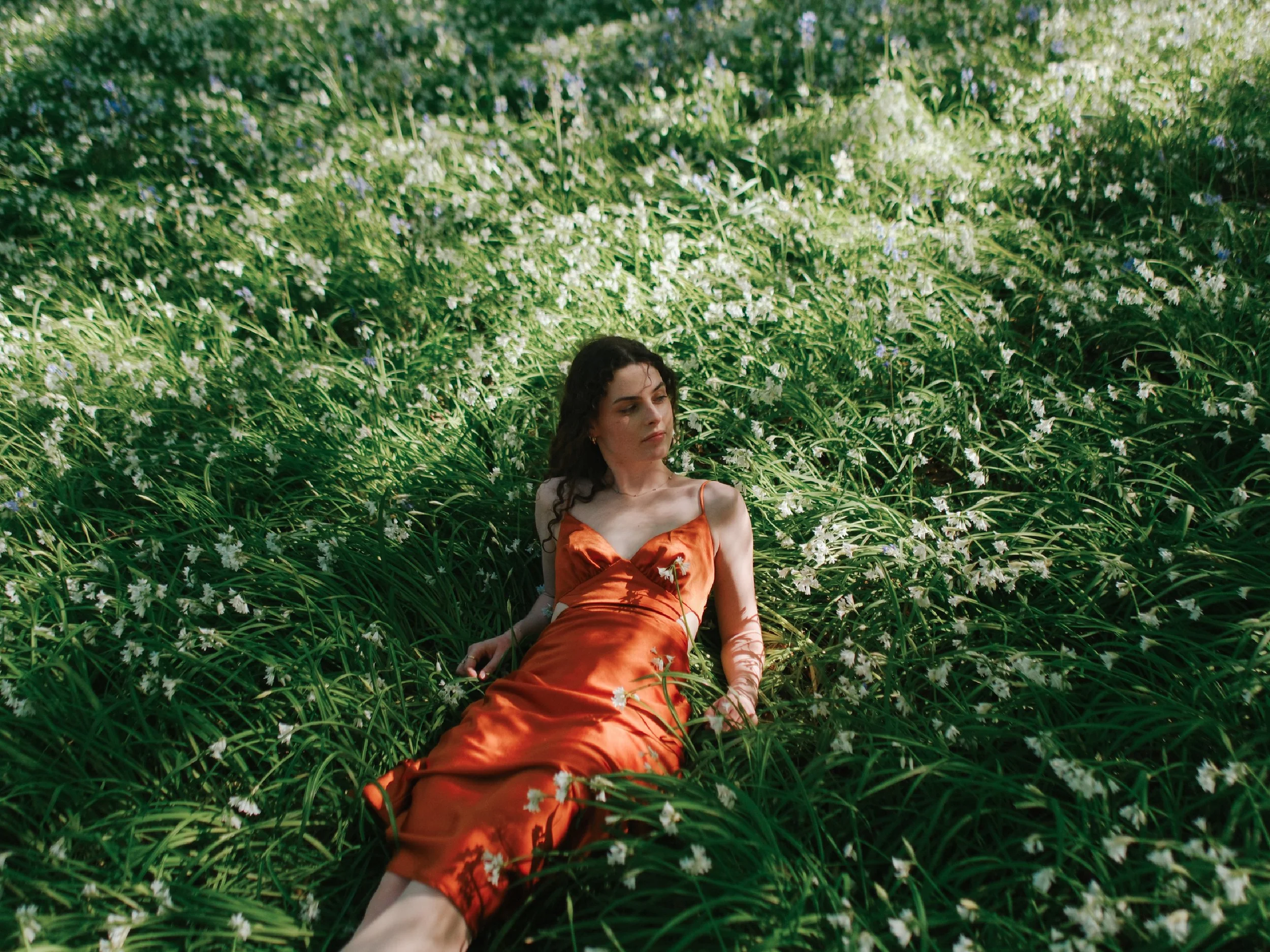 A woman in an orange dress is lying on green grass in a field of white flowers.