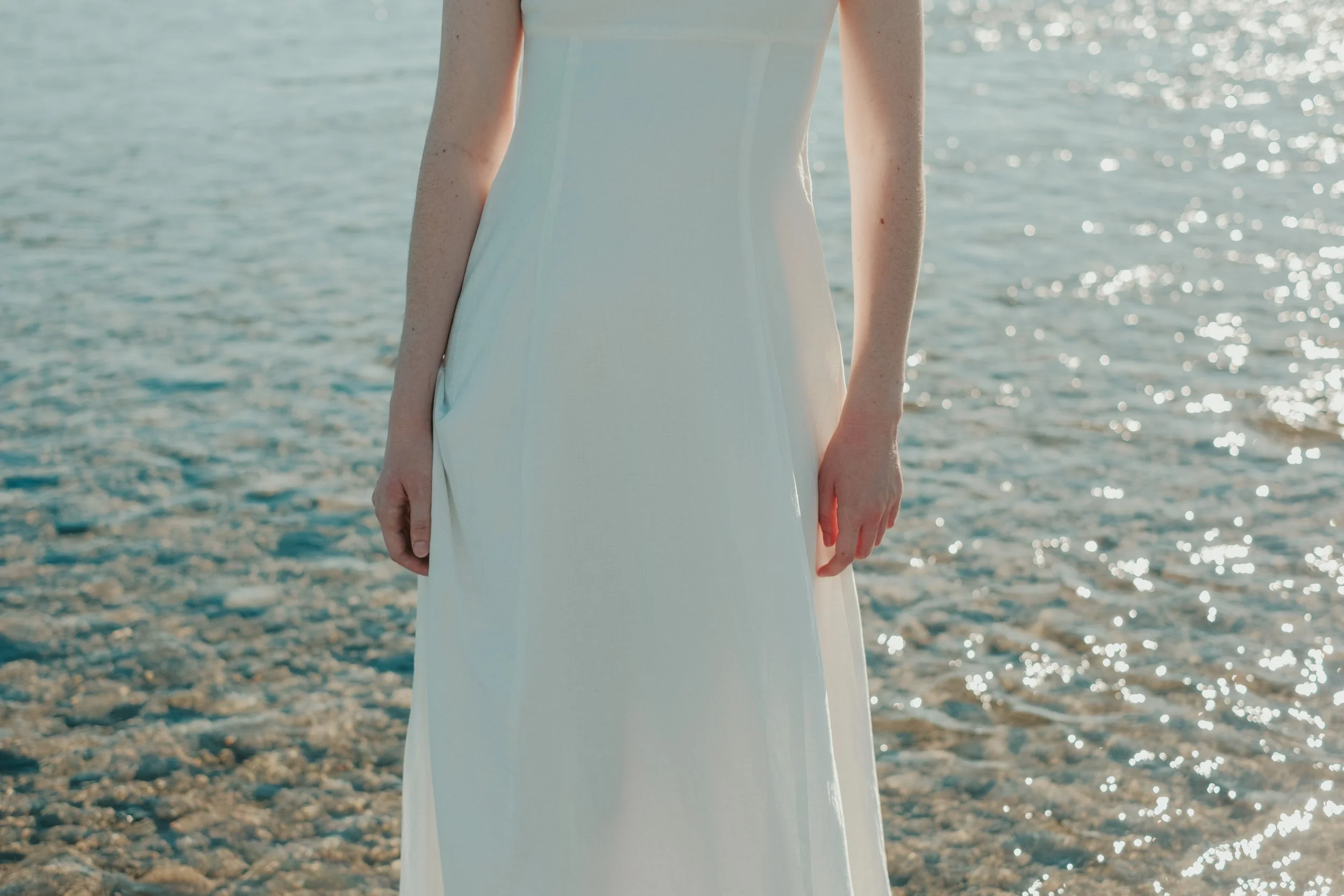 A woman in a white dress standing on a rocky beach near the water.