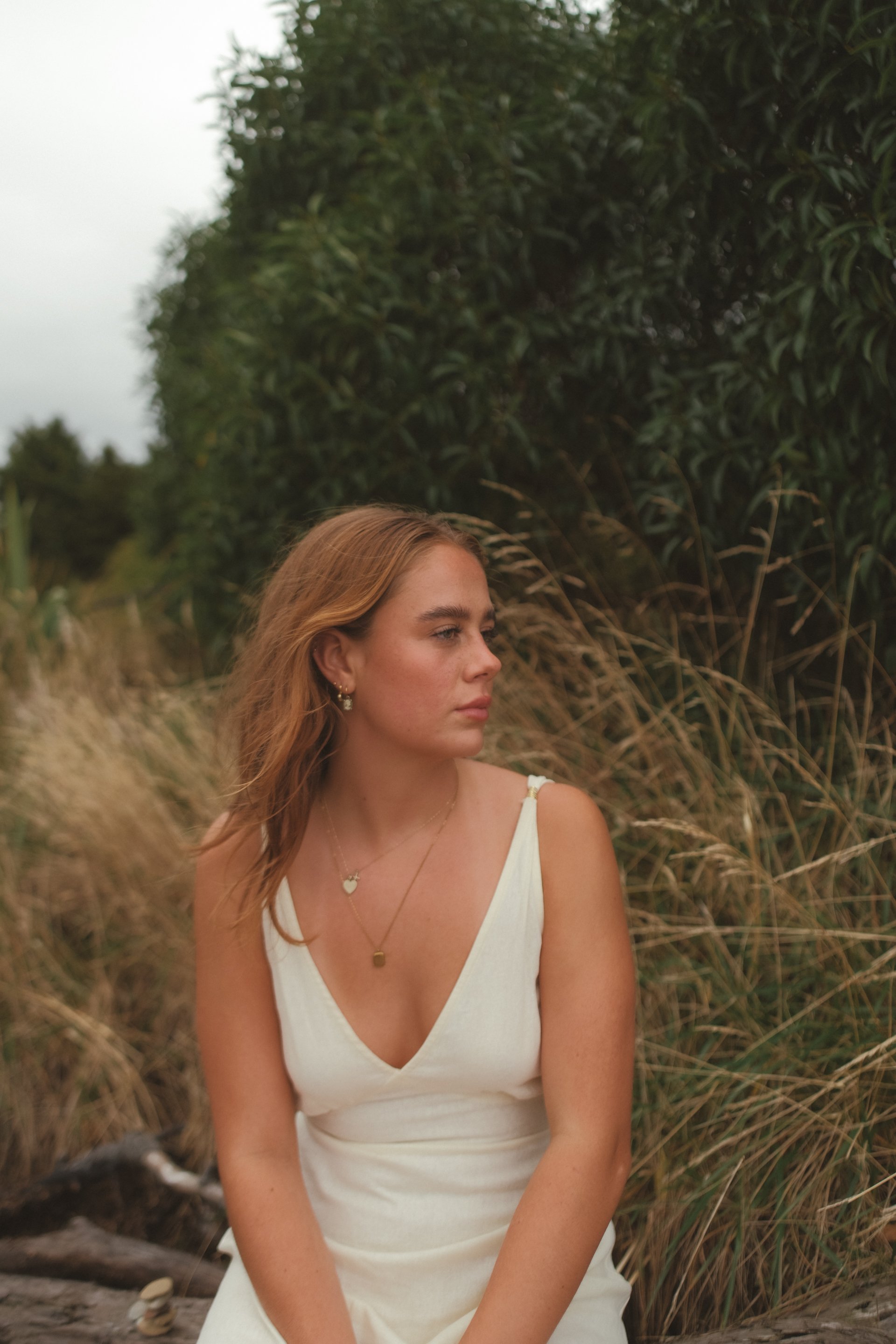 A young woman with light brown hair is sitting outdoors near tall grass and bushes, wearing a white sleeveless dress and gold jewelry, looking to the side.