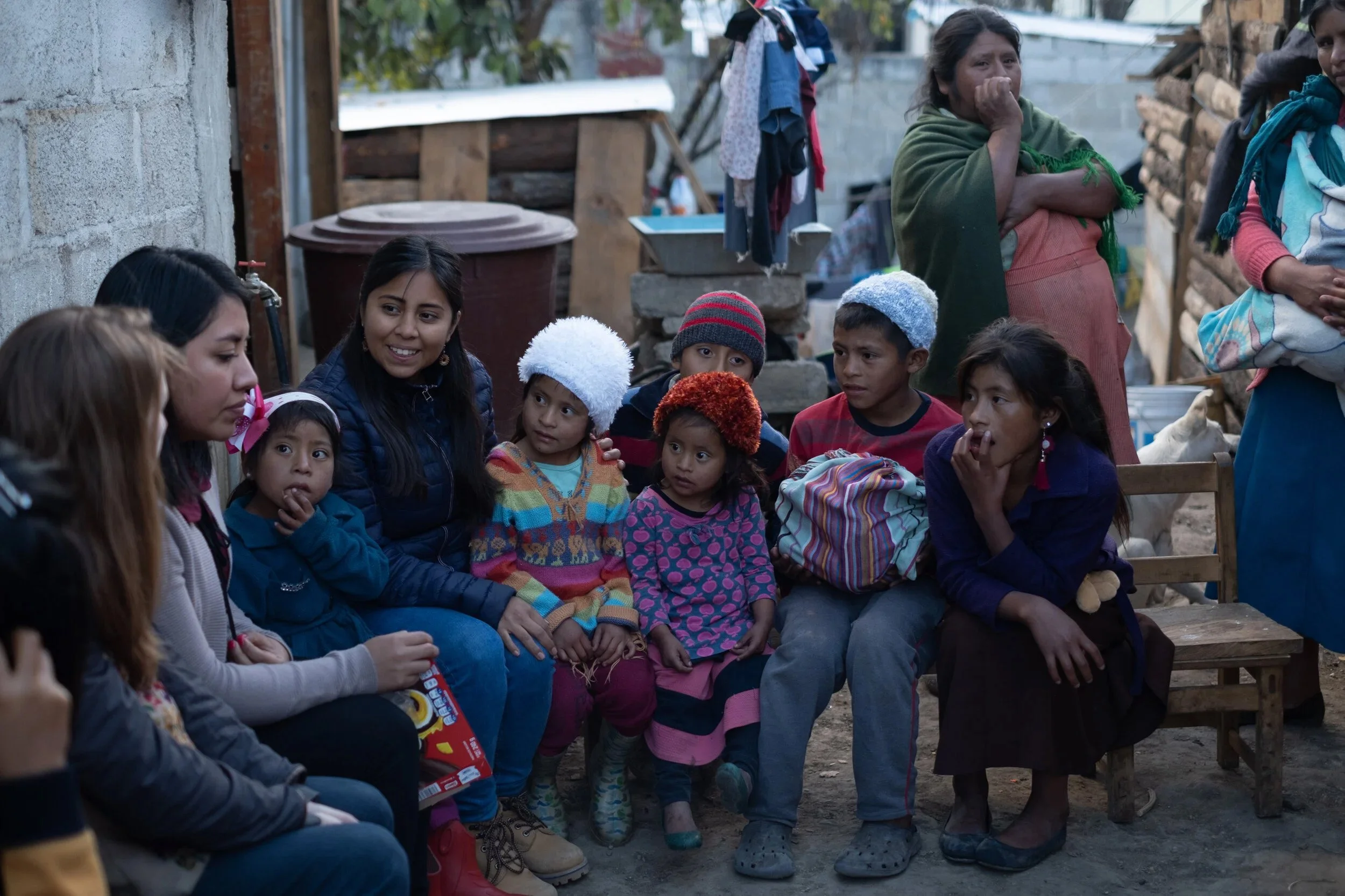 Group of children and women sitting outdoors in a semi-circle, engaging in a conversation. Some children wear colorful sweaters and hats, while the women are dressed in layered clothing. The setting appears to be in a rural or underdeveloped area with makeshift structures in the background.