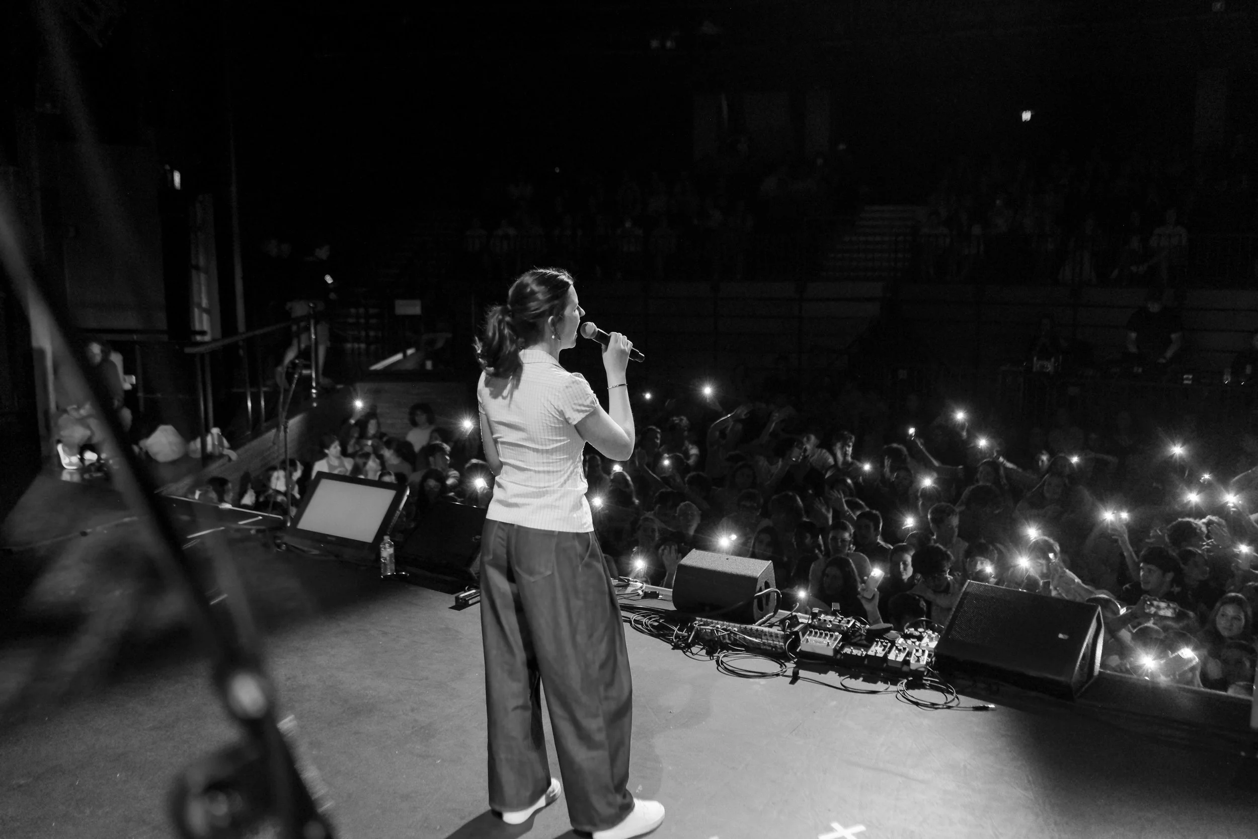A woman singing on stage with a microphone, facing a crowd with lights in their phones, in a concert setting.
