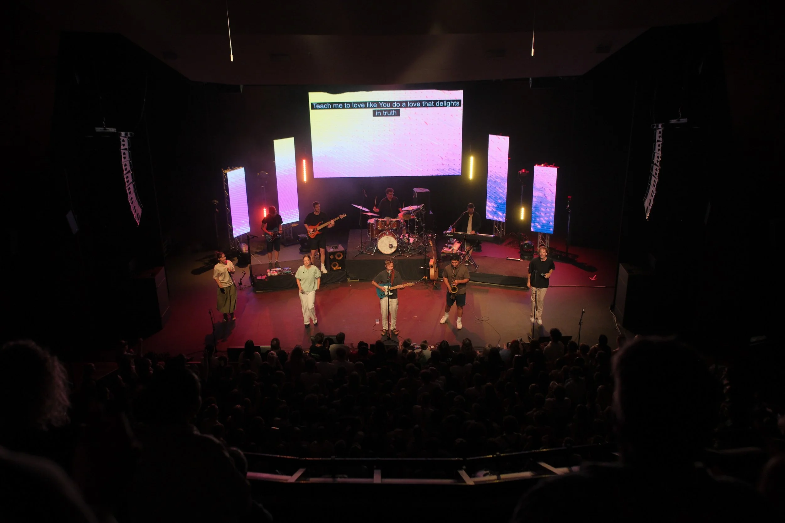 A live band performing on stage with seven musicians, colorful LED screens, and audience seated in a dark theater.