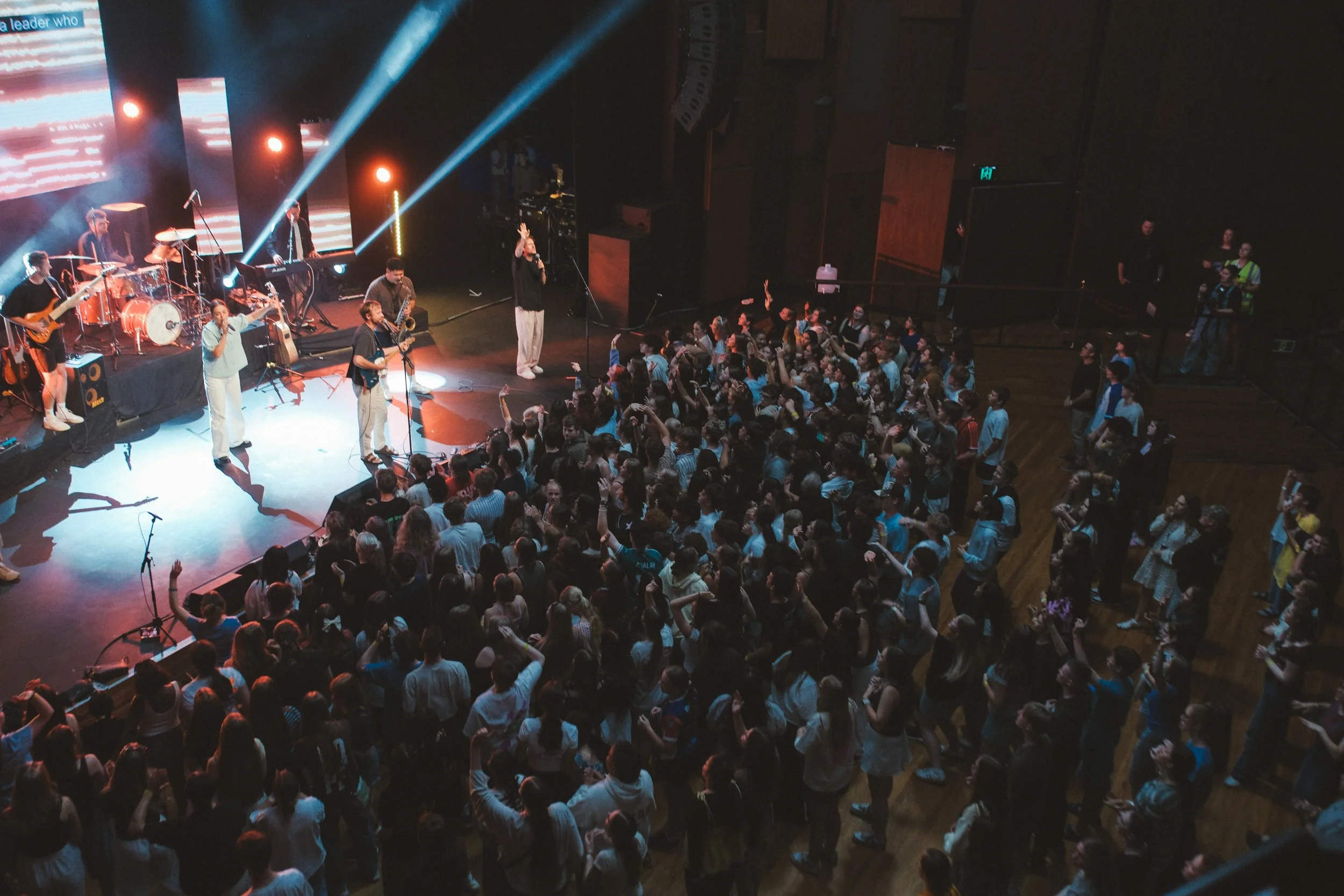 A band performs on stage with instruments and microphones as a large crowd dances and enjoys the concert in an indoor venue with wooden floors.