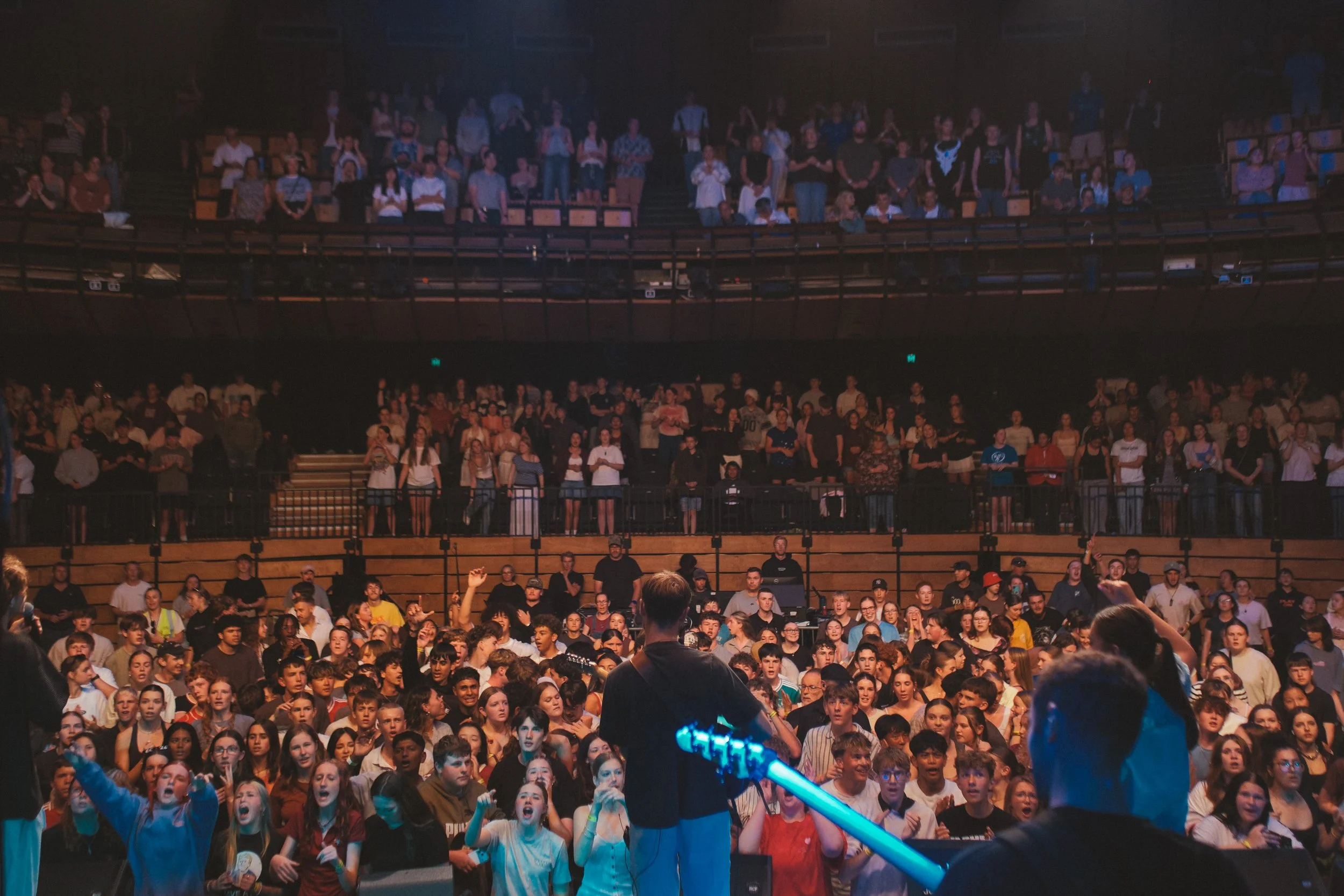Crowd of young people at a concert or event in a large indoor venue with multiple levels of seating, some standing and some watching the stage where a musician is performing.