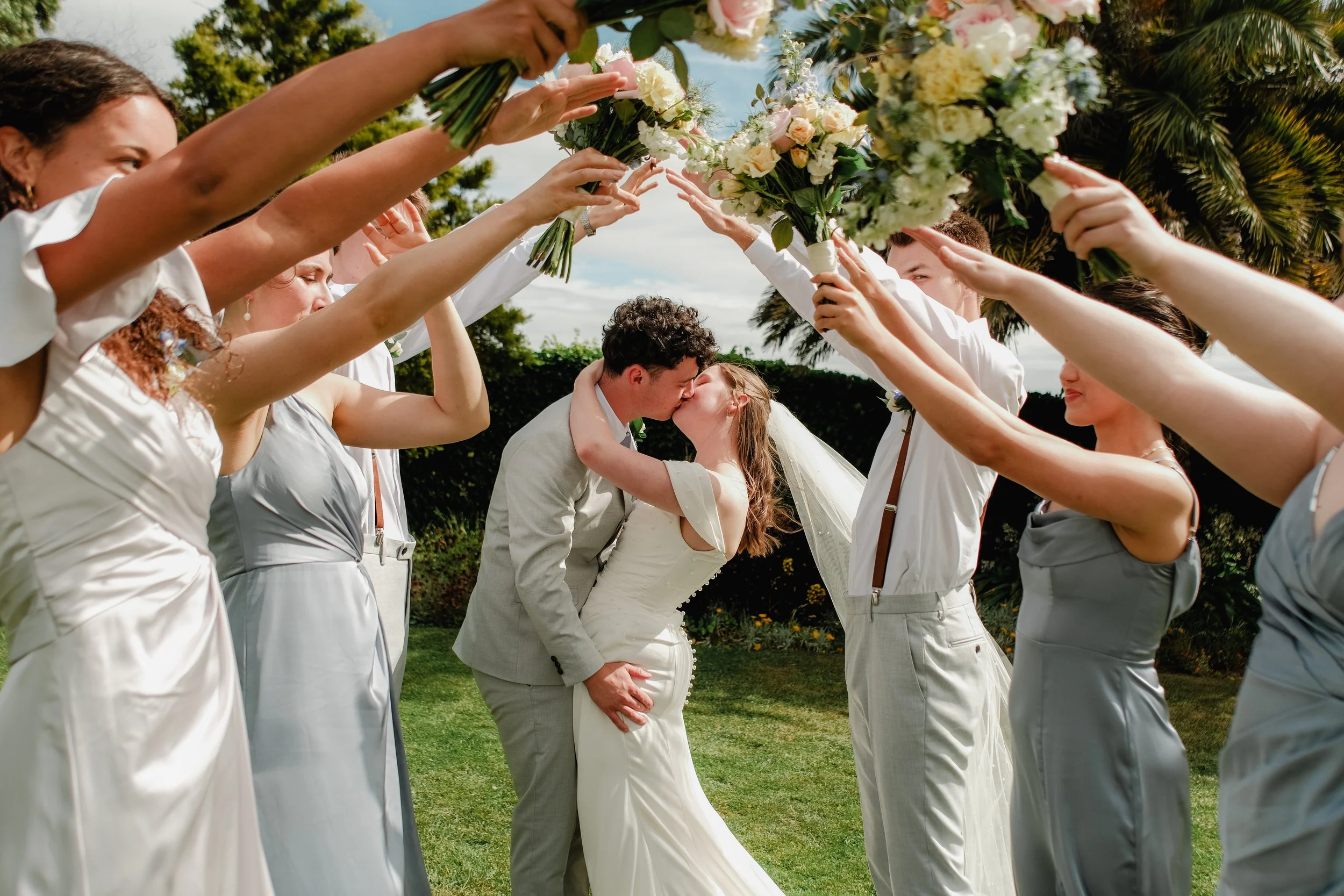 Bride and groom kissing under a floral arch, surrounded by bridesmaids and groomsmen holding bouquets and dressed in light-colored clothes, outdoors with greenery and palm trees.
