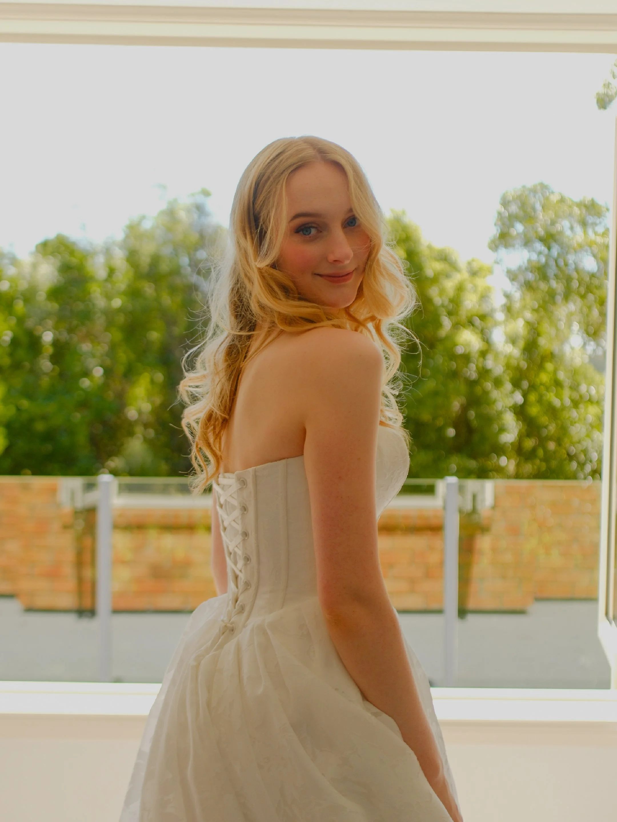 A young woman in a white dress standing by a window with a view of trees outside.