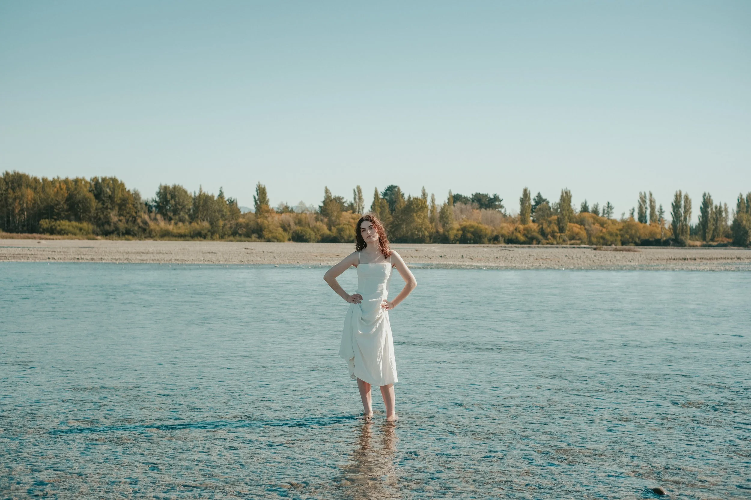 Woman in a white dress standing in shallow water with trees in the background.