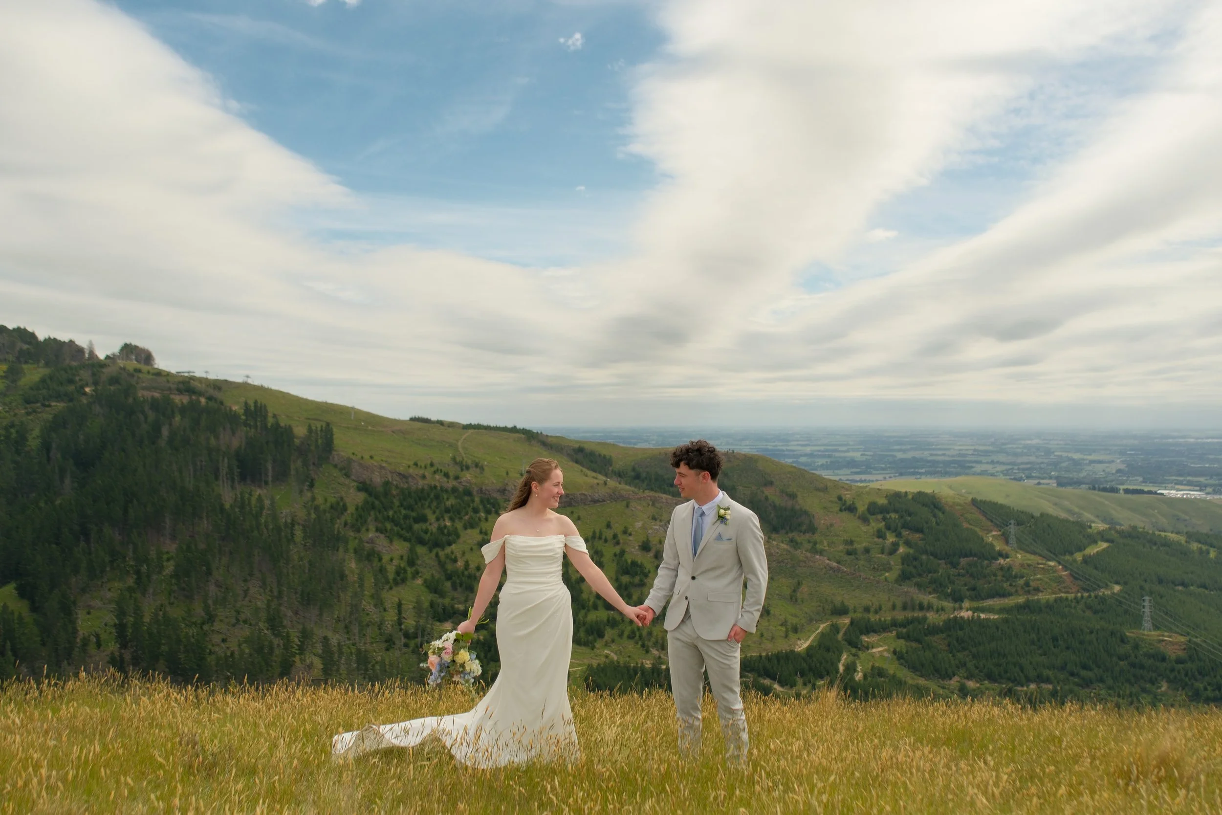 A bride and groom holding hands in a grassy field with rolling green hills and a cloudy sky in the background.