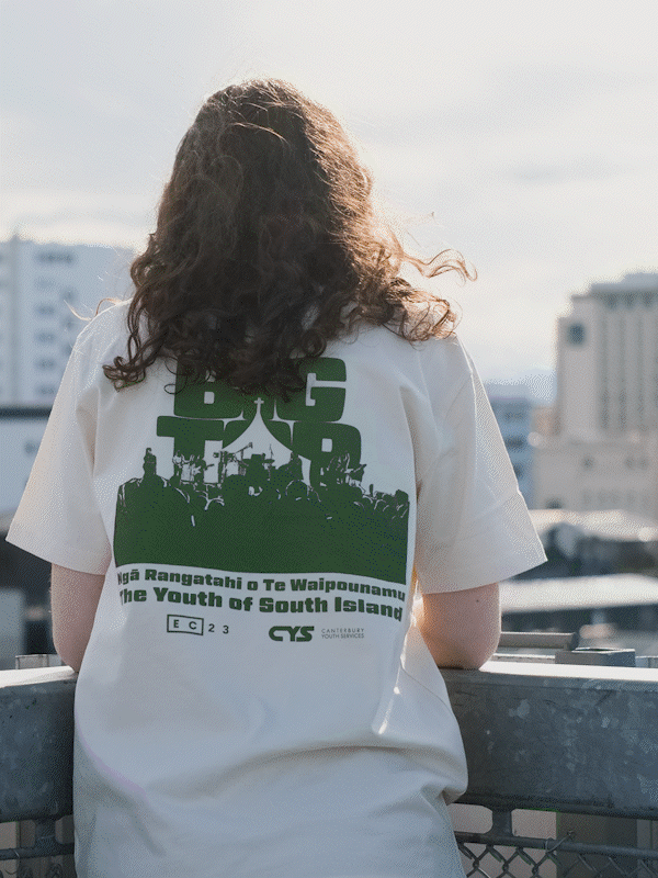 Person with long curly hair wearing a white t-shirt with green text and graphics, standing on a rooftop with a cityscape in the background.