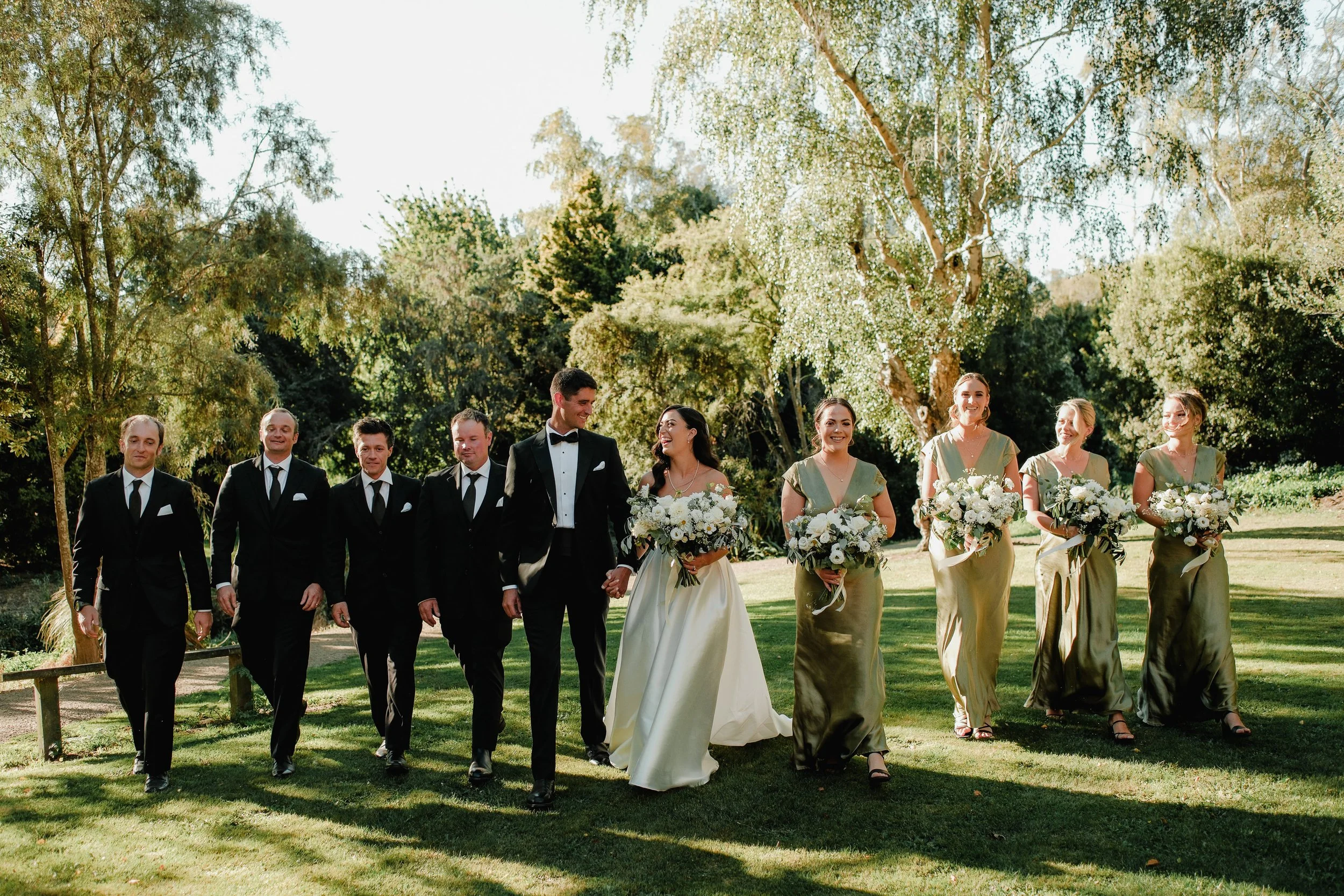 A wedding party walking outdoors on a sunny day, consisting of the bride and groom in the center, with bridesmaids and groomsmen on either side, all holding bouquets and dressed in formal attire, surrounded by green trees and grass.