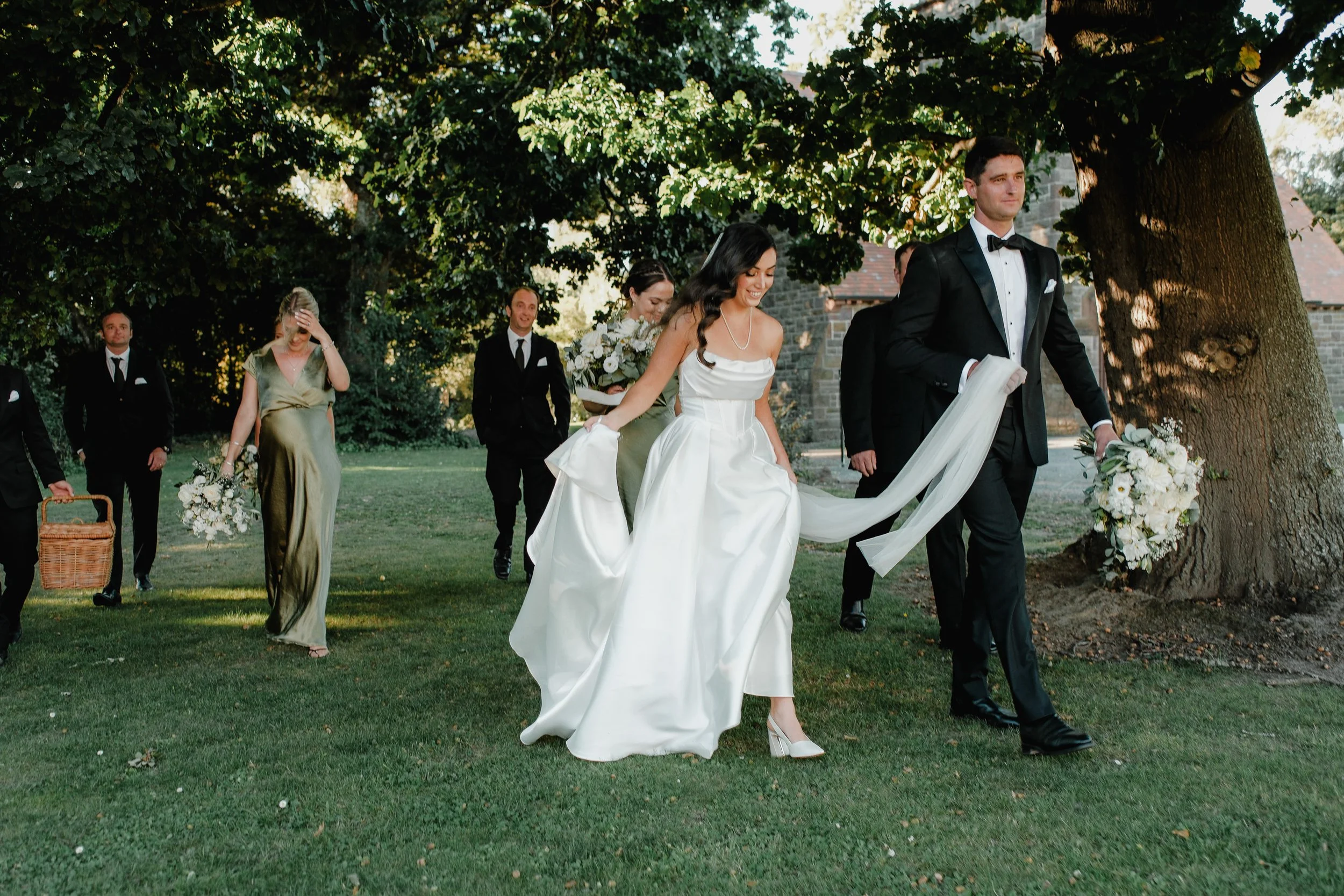 Wedding party walking outdoors under a large tree, including a bride in a white gown and a groom in a black tuxedo, with bridesmaids and groomsmen dressed in formal attire, holding bouquets and walking on grass.