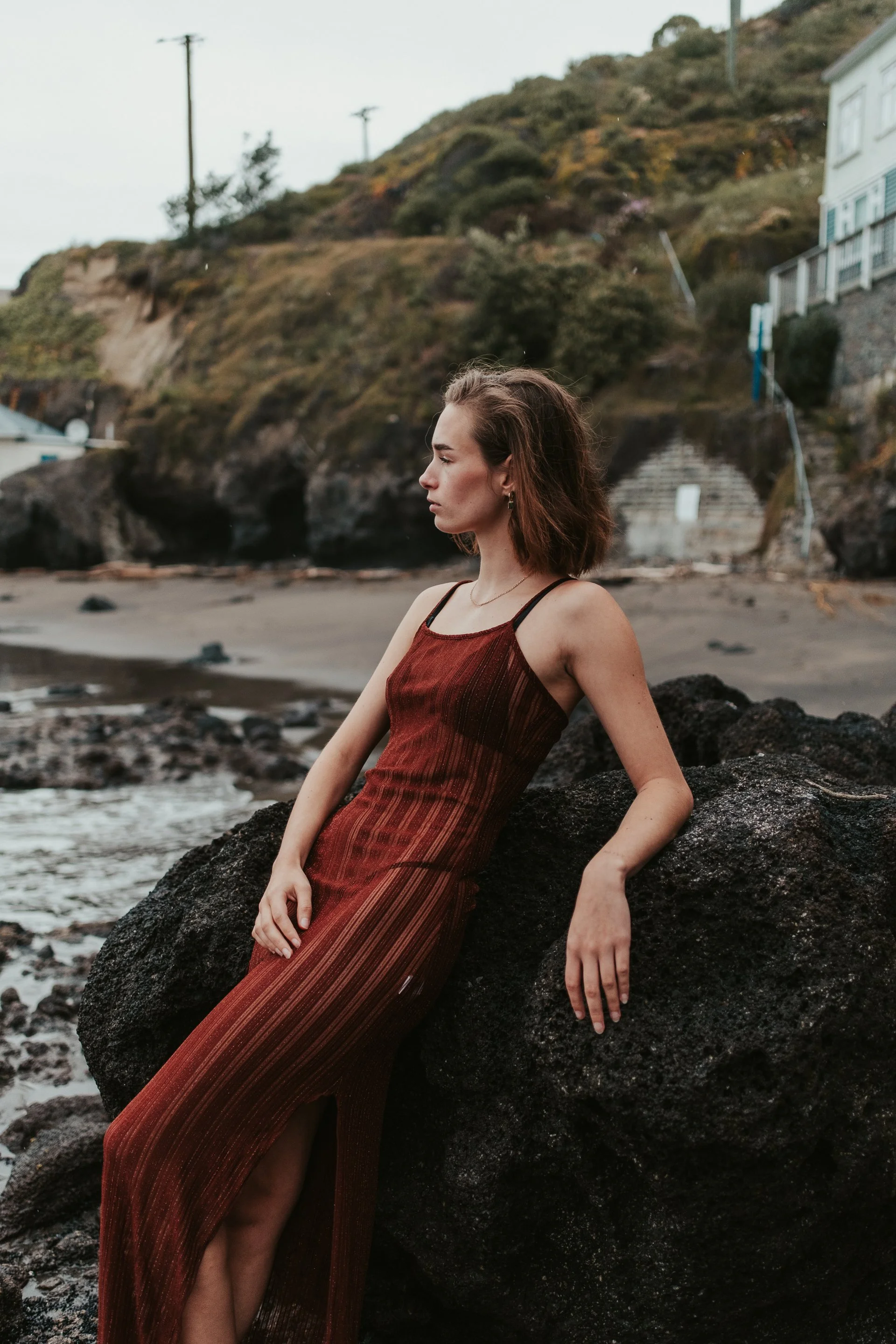 A young woman in a long, sleeveless, rust-colored dress with black straps sitting on a large black rock by the beach, with hillside homes and stairs in the background.