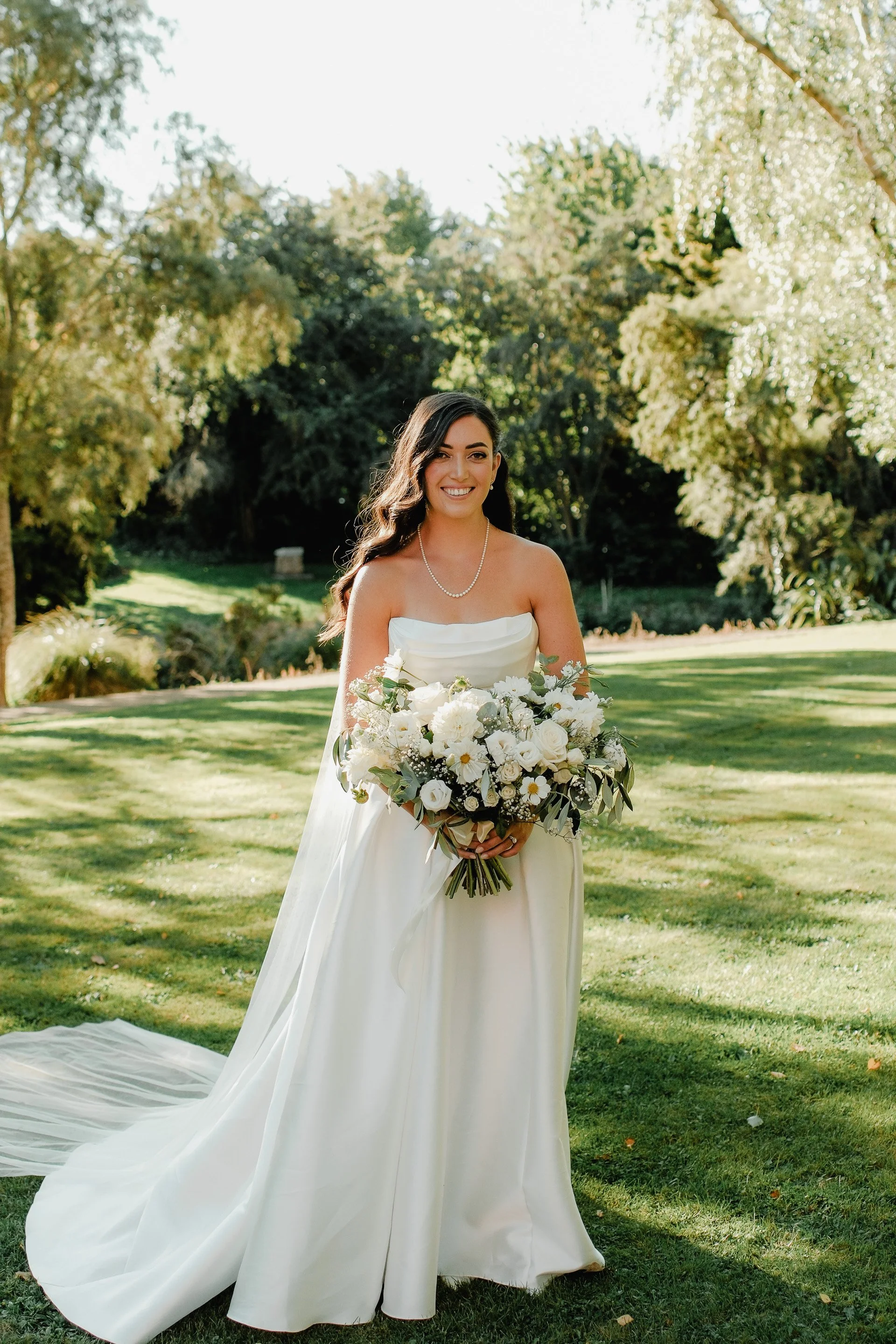 A bride in a strapless white wedding gown holding a bouquet of white flowers outdoors on a sunny day, with trees and green grass in the background.