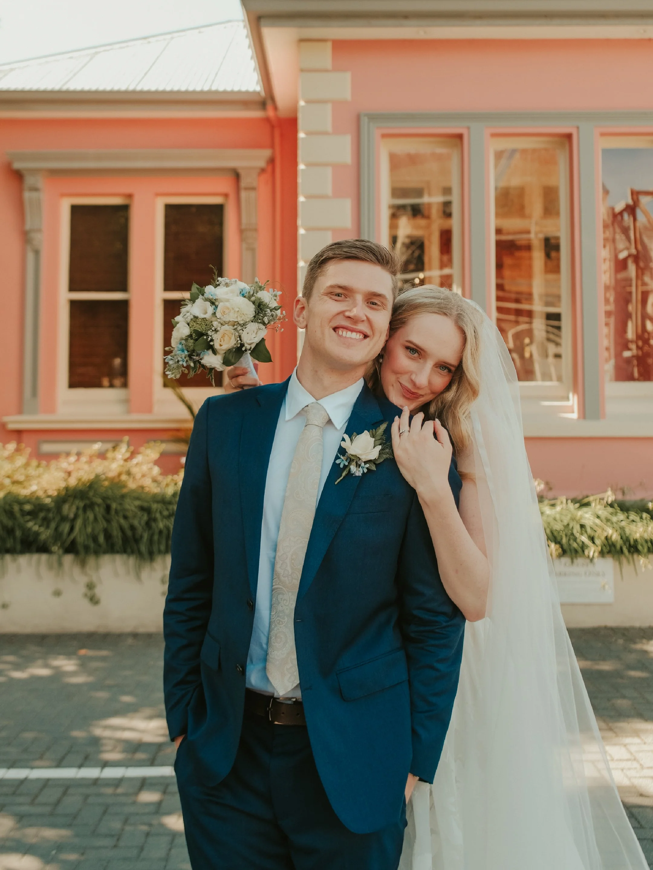 A smiling bride and groom posing outdoors in front of a pink building, with the bride gently resting her head on the groom's shoulder, holding a bouquet of flowers.